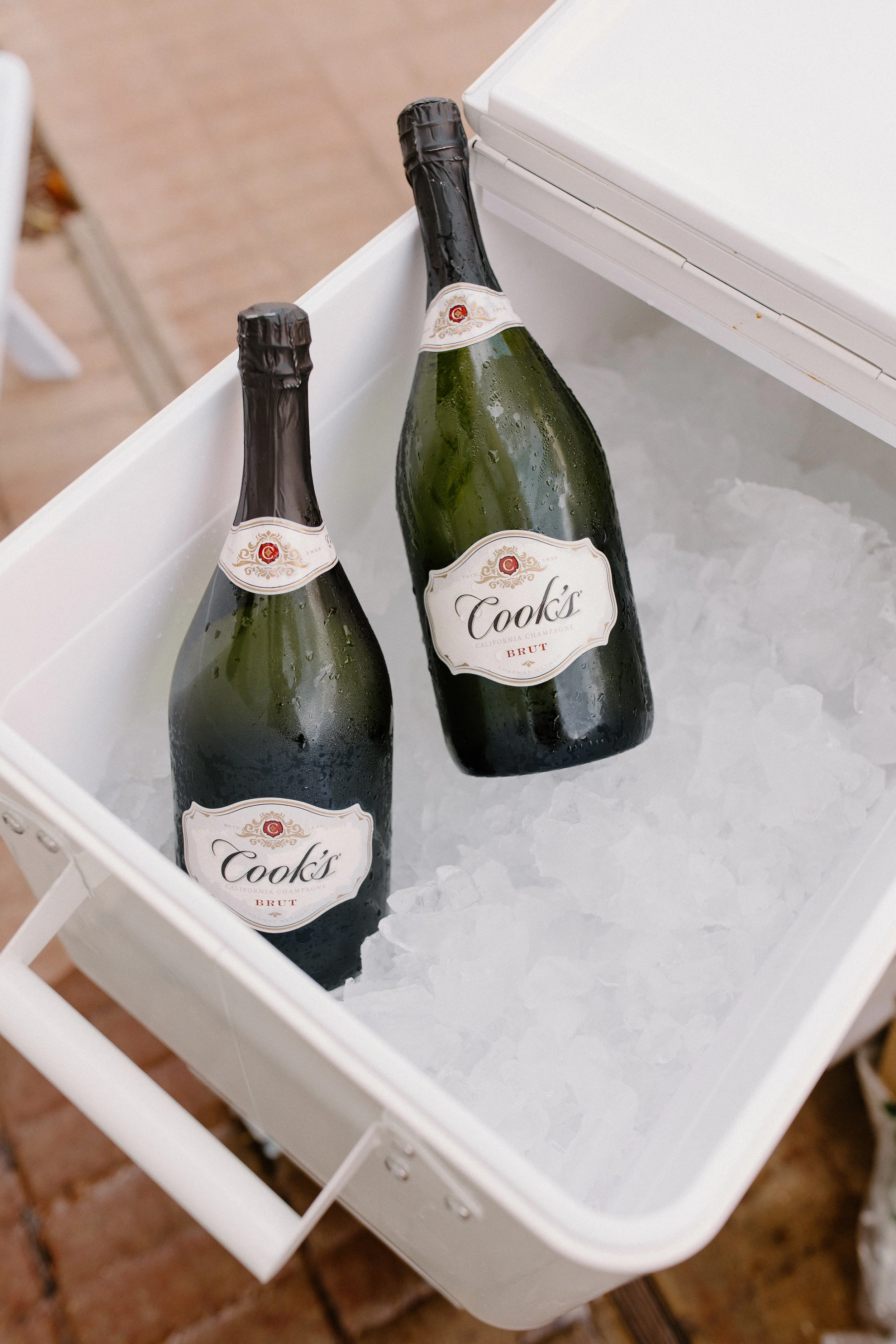 Two bottles of Cook's California Champagne Brut in an ice-filled cooler with a white exterior, placed on a brick surface.