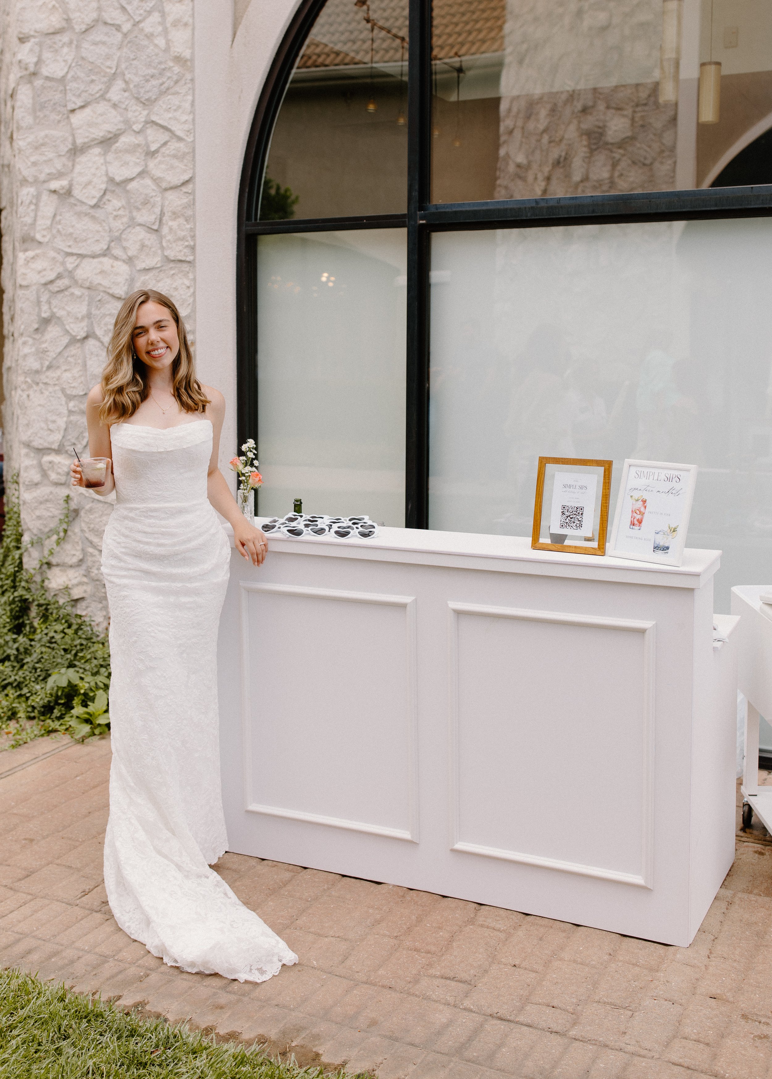 A woman in a white lace wedding dress with a strapless neckline stands outdoors next to a white bar or drink stand. She is holding a drink in her right hand, smiling, and her hair is styled in loose waves. The stand has two framed signs, one with a QR code and the other with drink options, and a small flower arrangement. The background includes a stone wall, a large black-framed window, some greenery, and outdoor lighting.