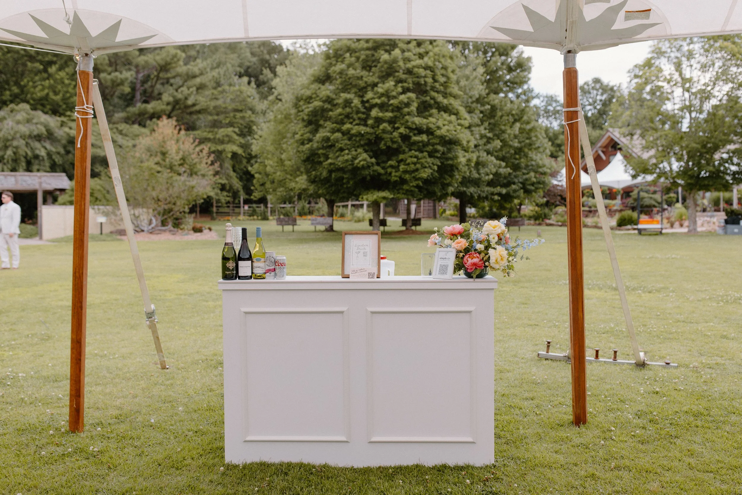 A small outdoor bar with wine bottles, a flower arrangement, and signs, set up under a canopy in a park or garden area with trees and benches in the background.