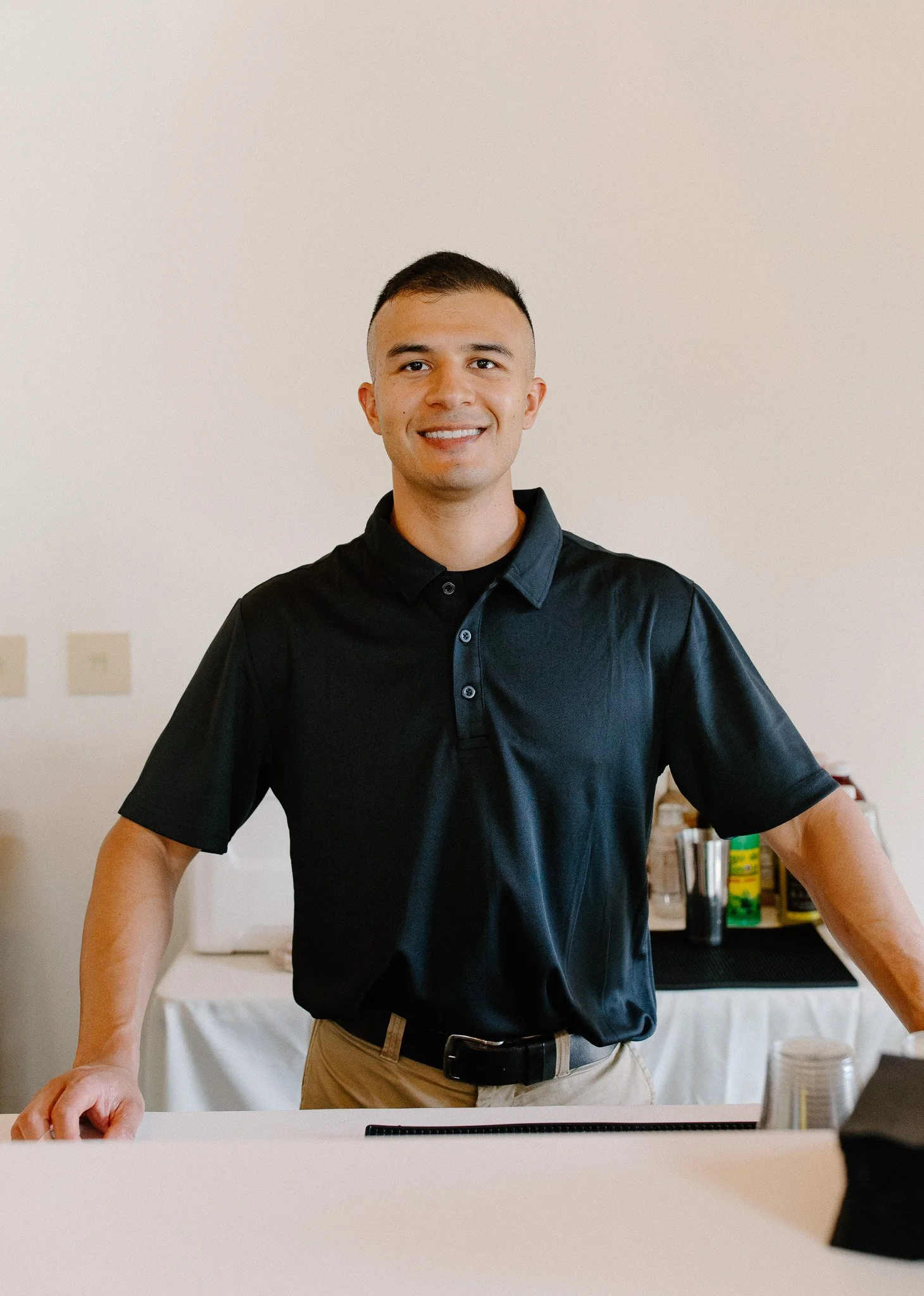 A young man with short dark hair, wearing a black polo shirt and khaki pants, smiling while standing behind a white counter in a kitchen or cafe setting.