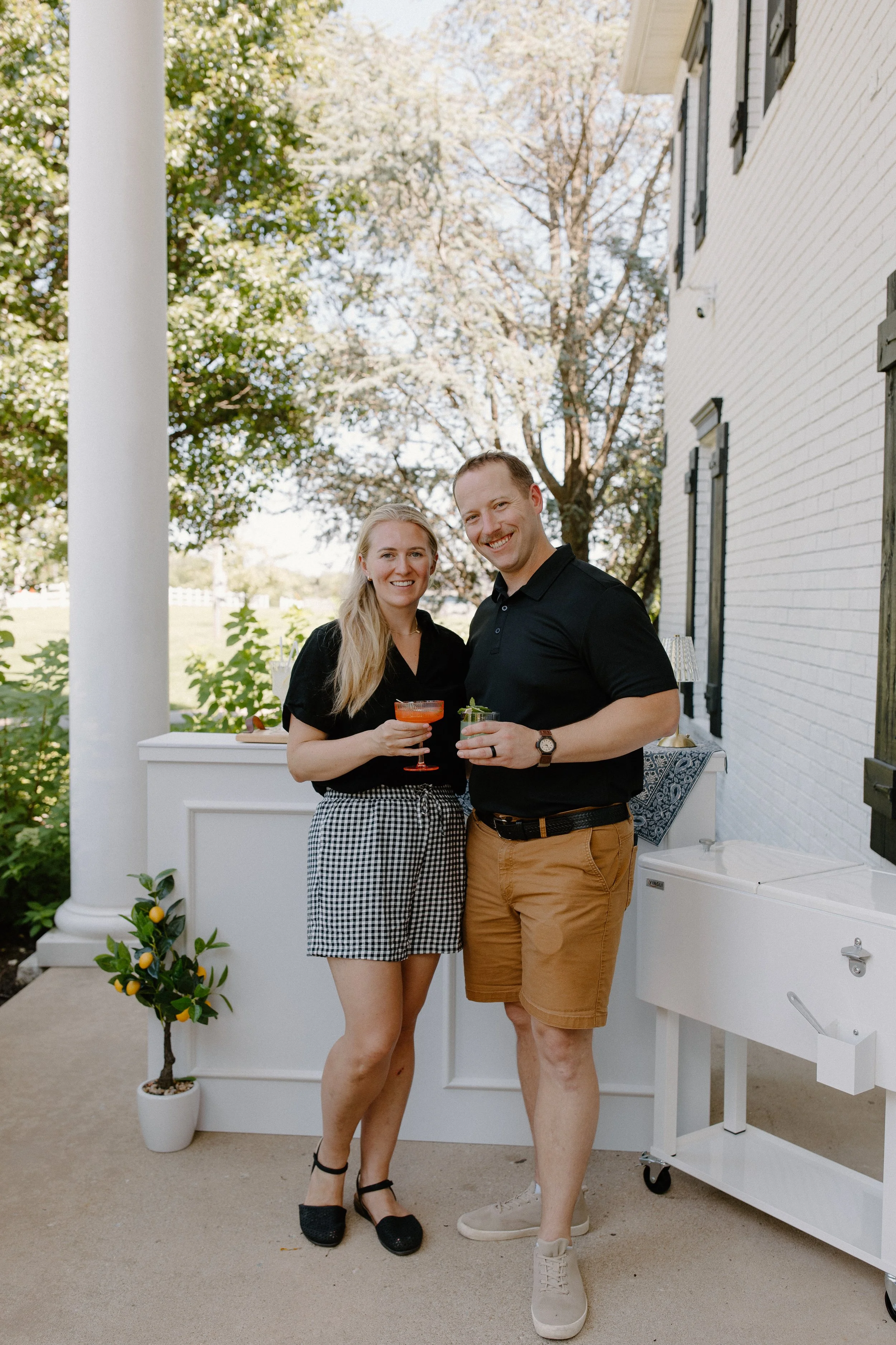 Two people standing on a porch, smiling and holding drinks, with a white brick house and trees with blooming branches in the background.