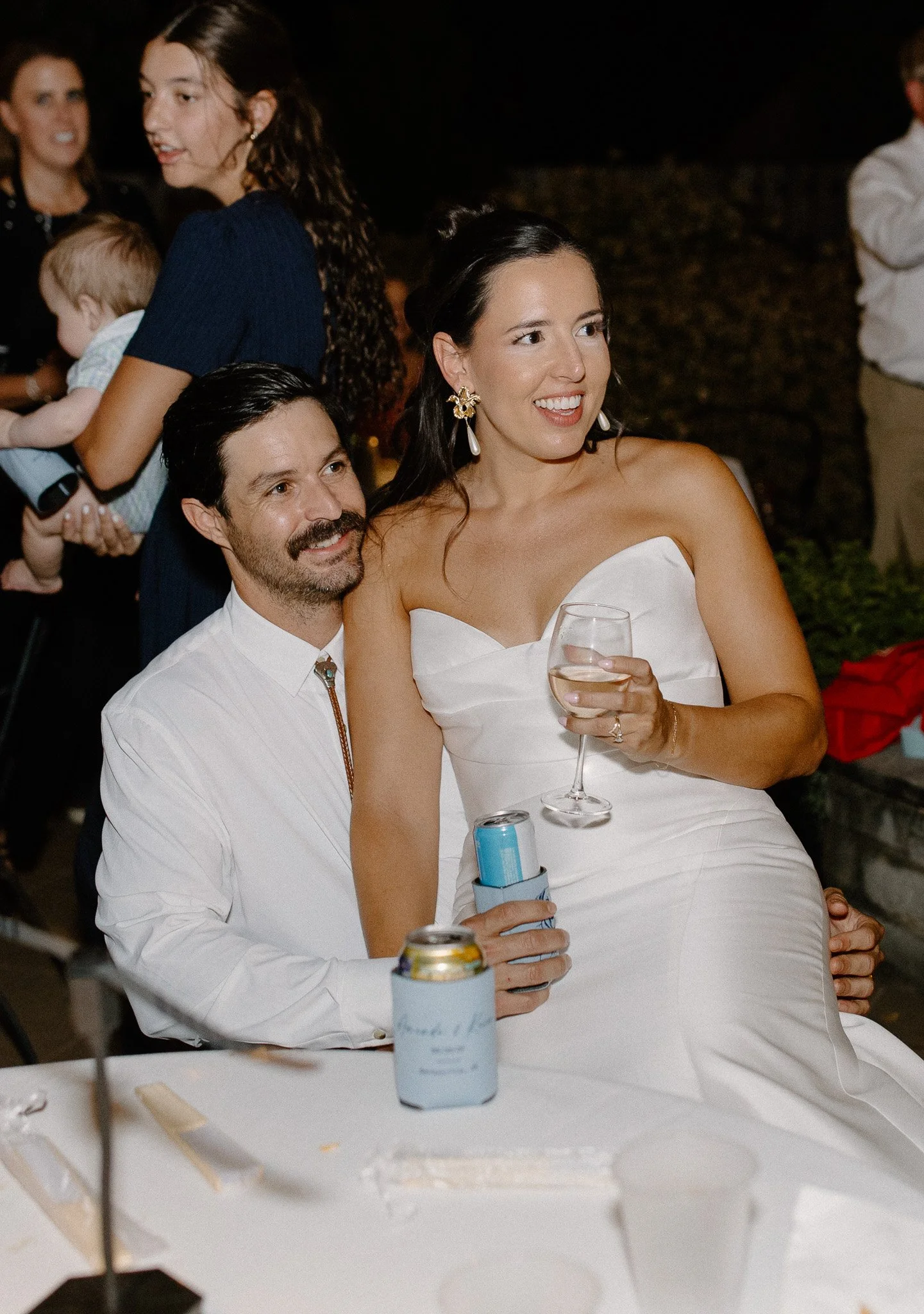 A couple at a celebration dinner, the woman wearing a white dress and holding a wine glass, the man with dark hair, a beard, and a white shirt. Other guests, including a woman holding a child, are in the background.
