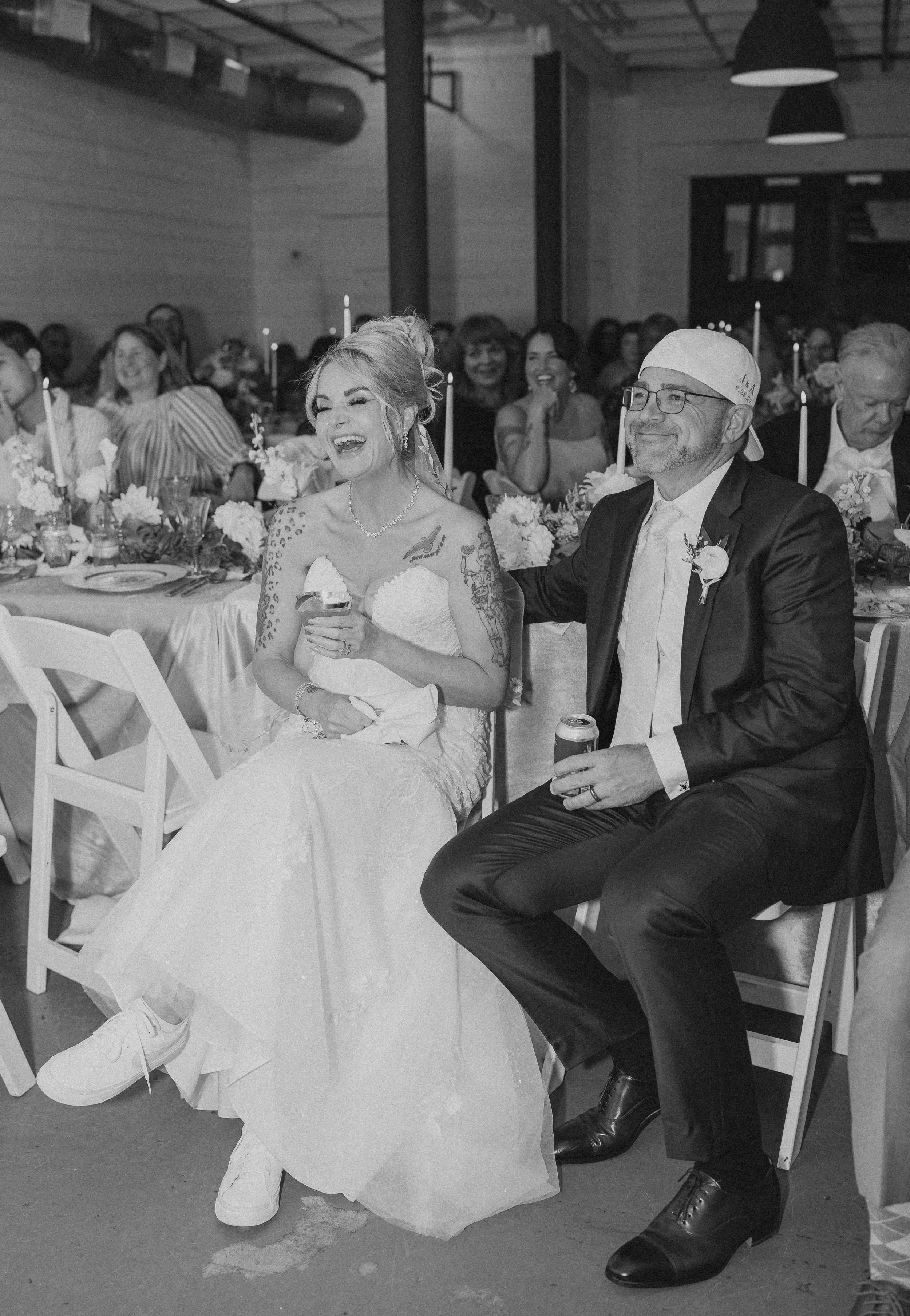 A bride and groom sitting at a wedding reception, laughing and holding drinks, surrounded by guests at decorated tables.