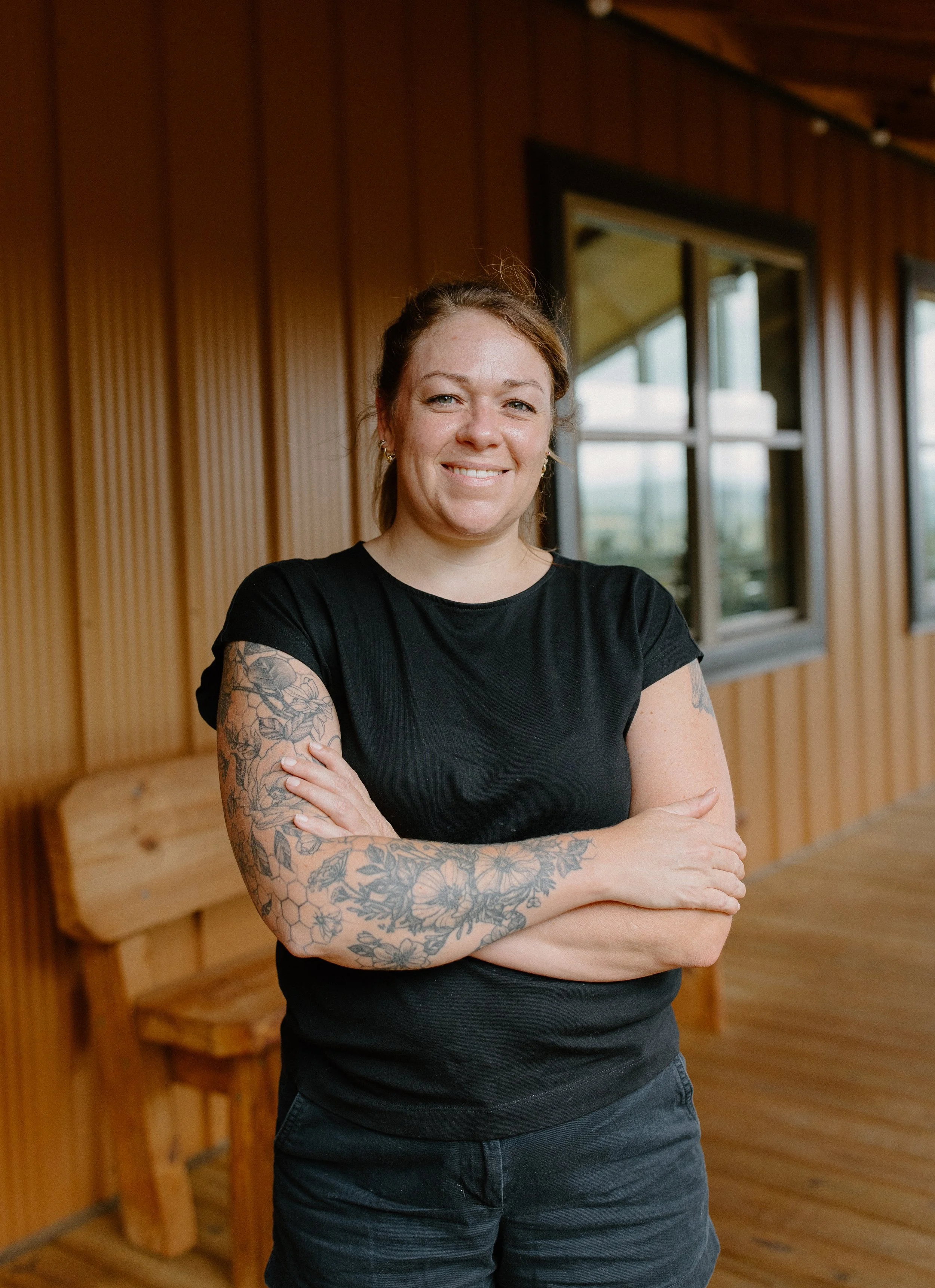 A woman with tattoos on her arms standing indoors next to a wooden bench and a window, smiling with her arms crossed.