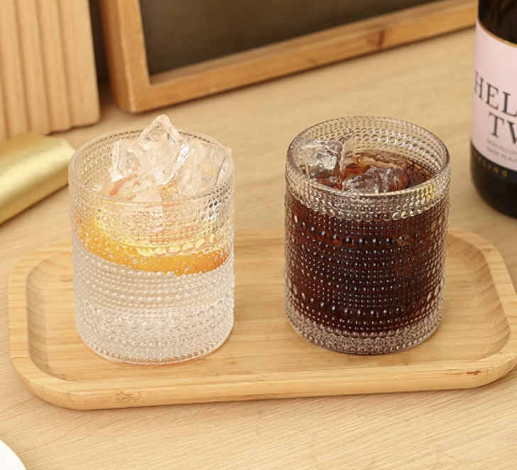 Two textured glass tumblers with ice, one with a lemon slice and the other with a dark soda, on a wooden tray, with a wine bottle partially visible in the background.