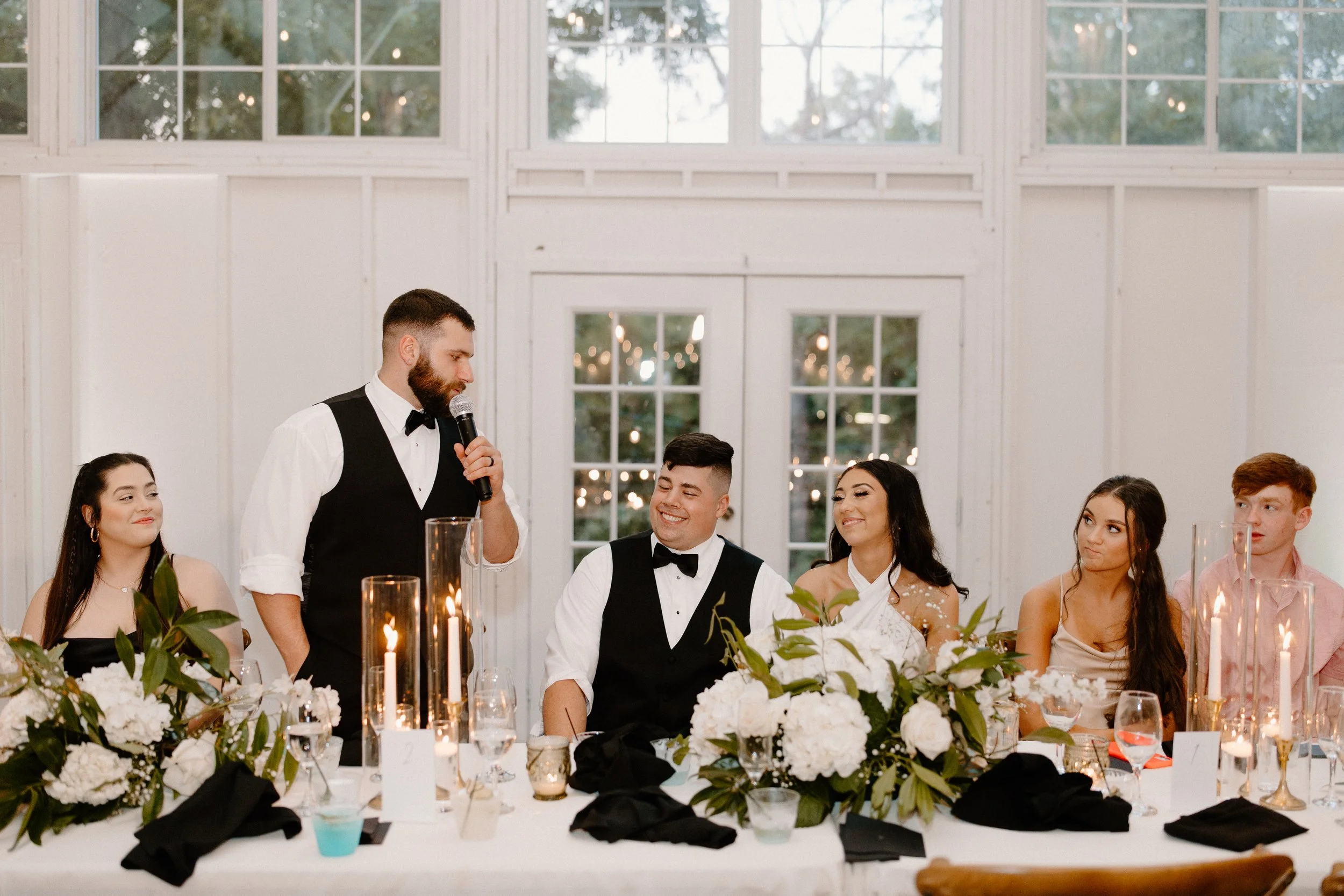 A man in a tuxedo and bow tie is giving a speech with a microphone at a wedding reception table, surrounded by smiling guests, elegant floral arrangements, and candles inside a venue with large windows showing outdoor trees.