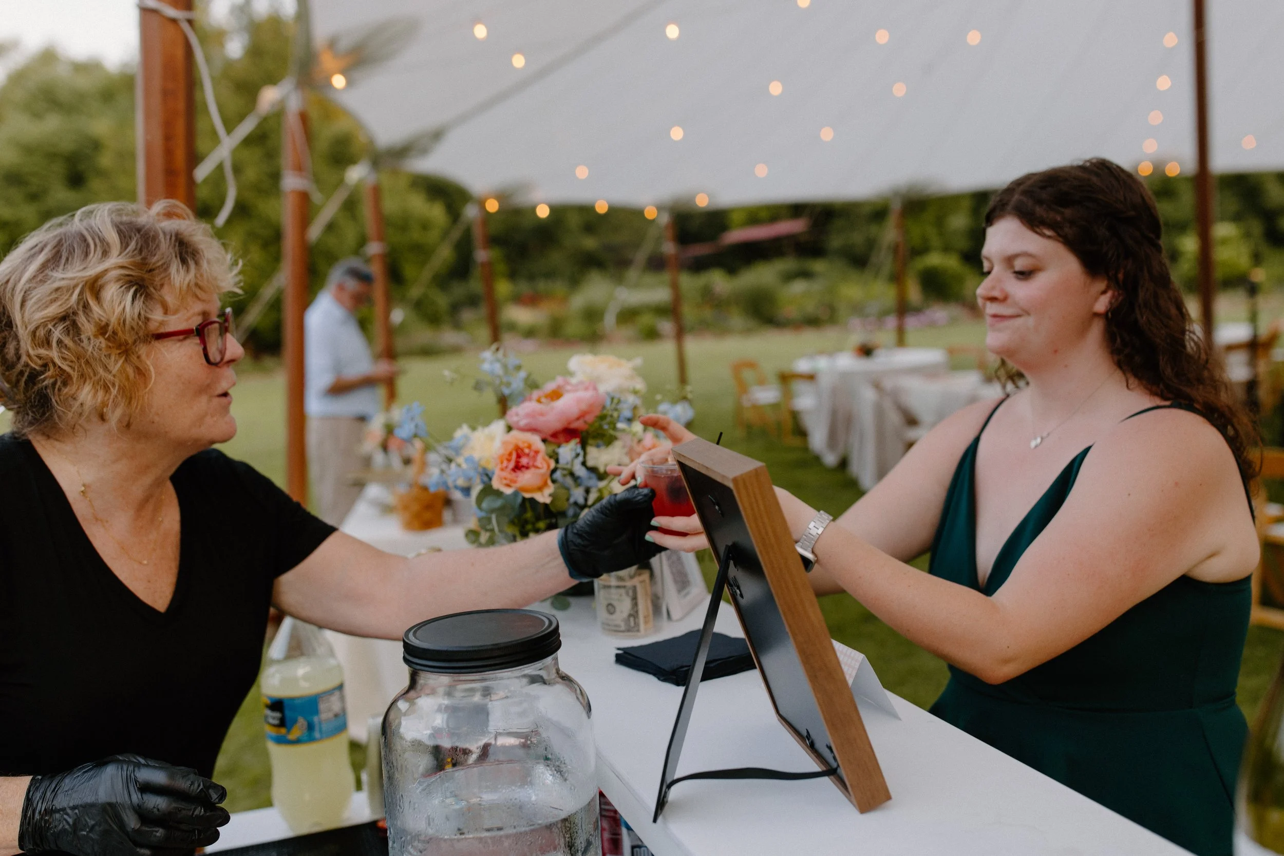 A woman with glasses and a woman with dark hair are exchanging a small item at a table outdoors during the evening with string lights. There is a floral arrangement and drinks on the table.