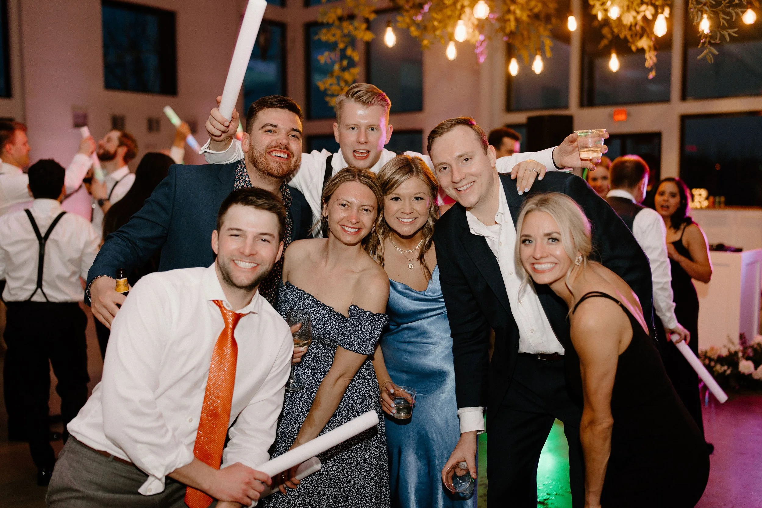 Group of friends at a party, smiling and posing for the photo. They are dressed in formal and semi-formal attire, holding drinks, with colorful lighting and a festive atmosphere in the background.