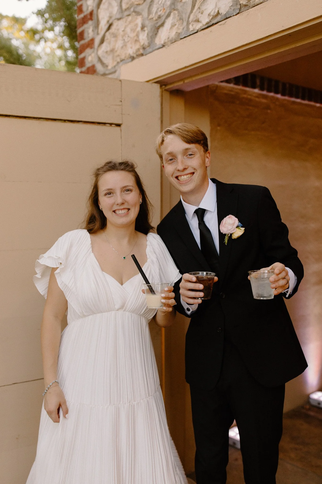 A woman in a white dress and a man in a black suit with a pink boutonniere are smiling and holding drinks at a social gathering.
