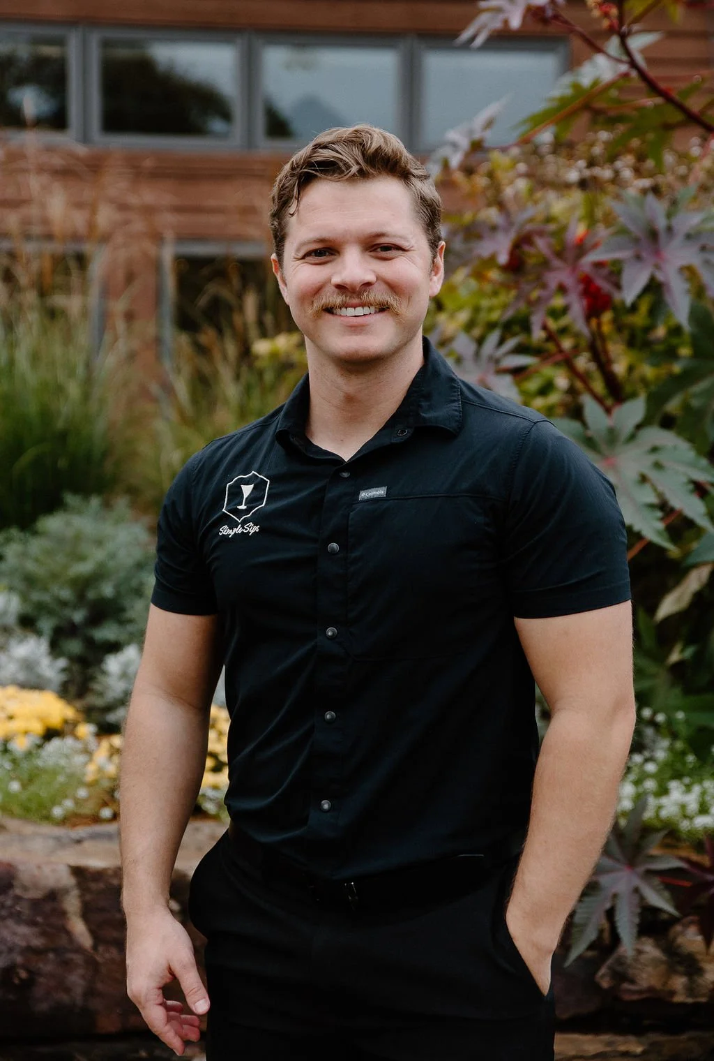 A man with light skin, wavy brown hair, and a moustache, smiling and wearing a black short-sleeved button-up shirt, standing outdoors in front of plants and a building.
