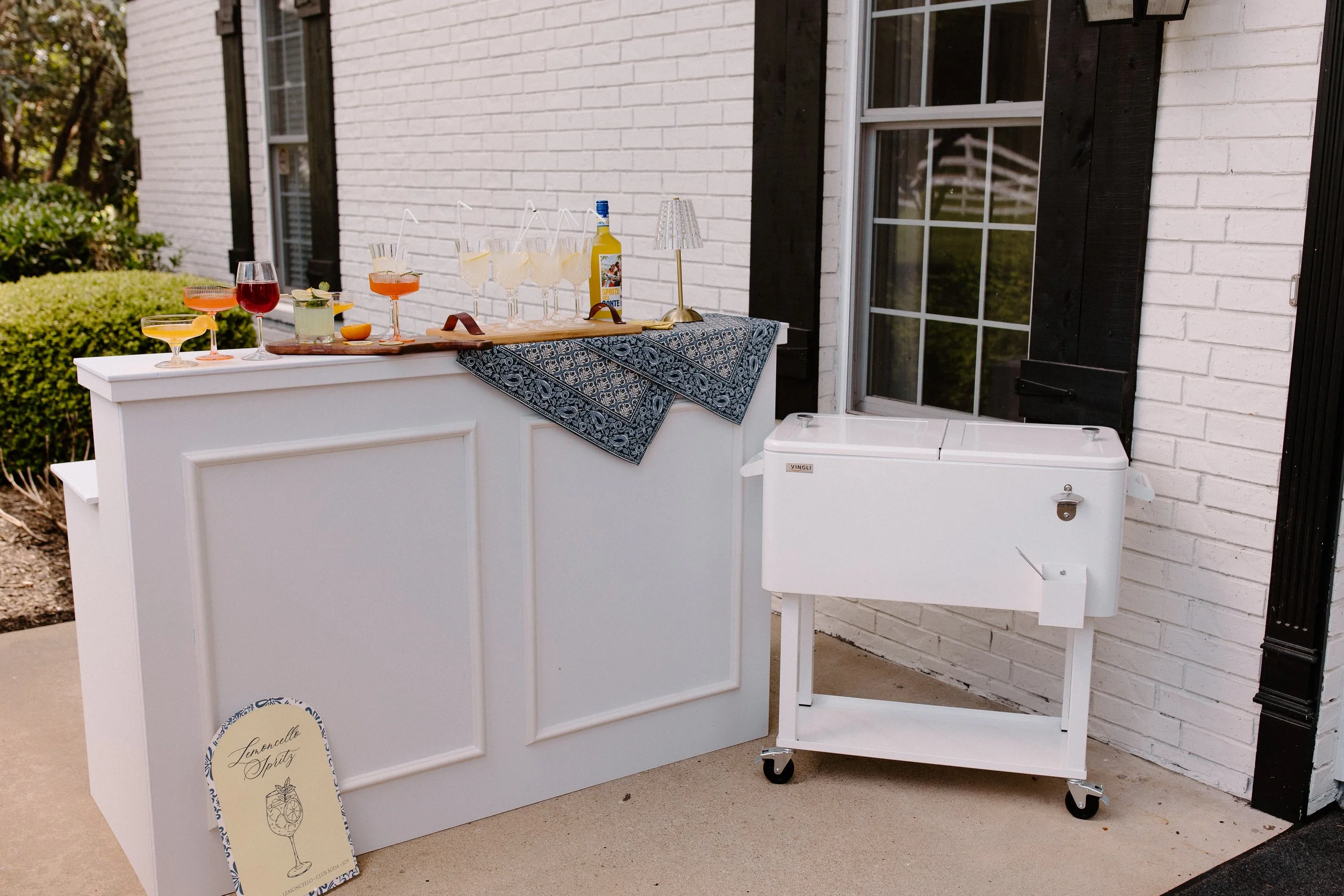 Outdoor beverage station with various cocktails and a lemon-flavored drink, a bubbly bottle, a decorative table lamp, and a sign that reads 'Lemongrass Spritz' outside a house.