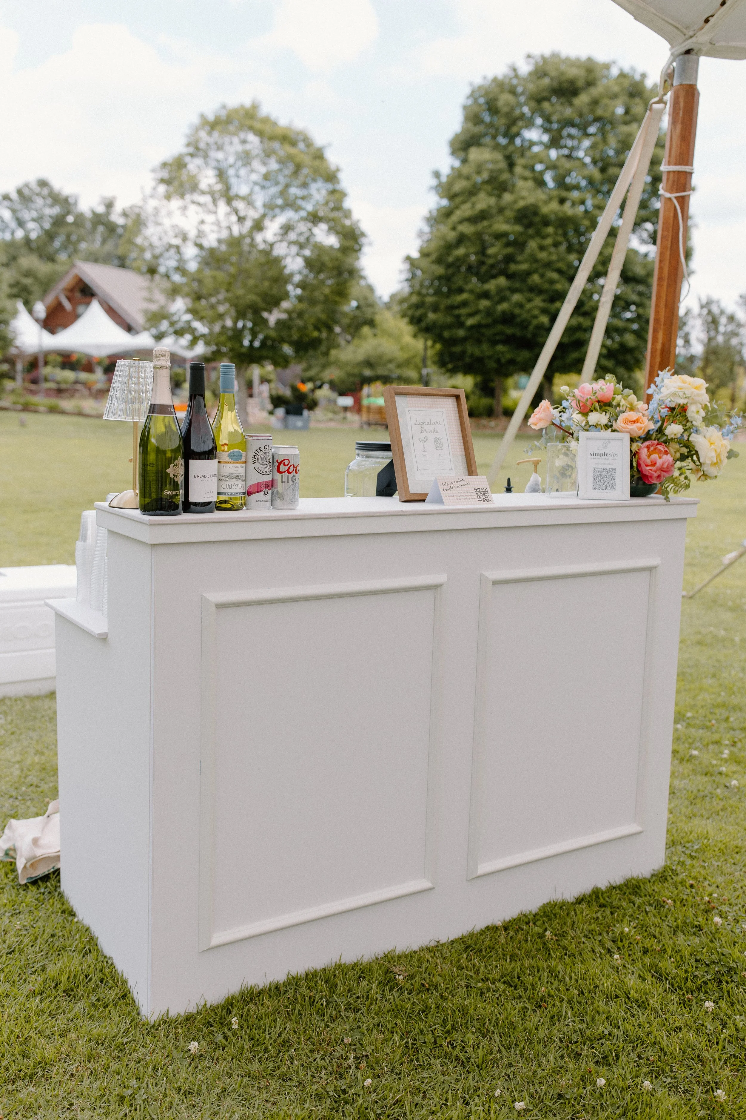 A white outdoor bar with bottles of wine, canned beverages, flower arrangements, and decorative signage, set up in a grassy area with trees and tents in the background for an outdoor event.