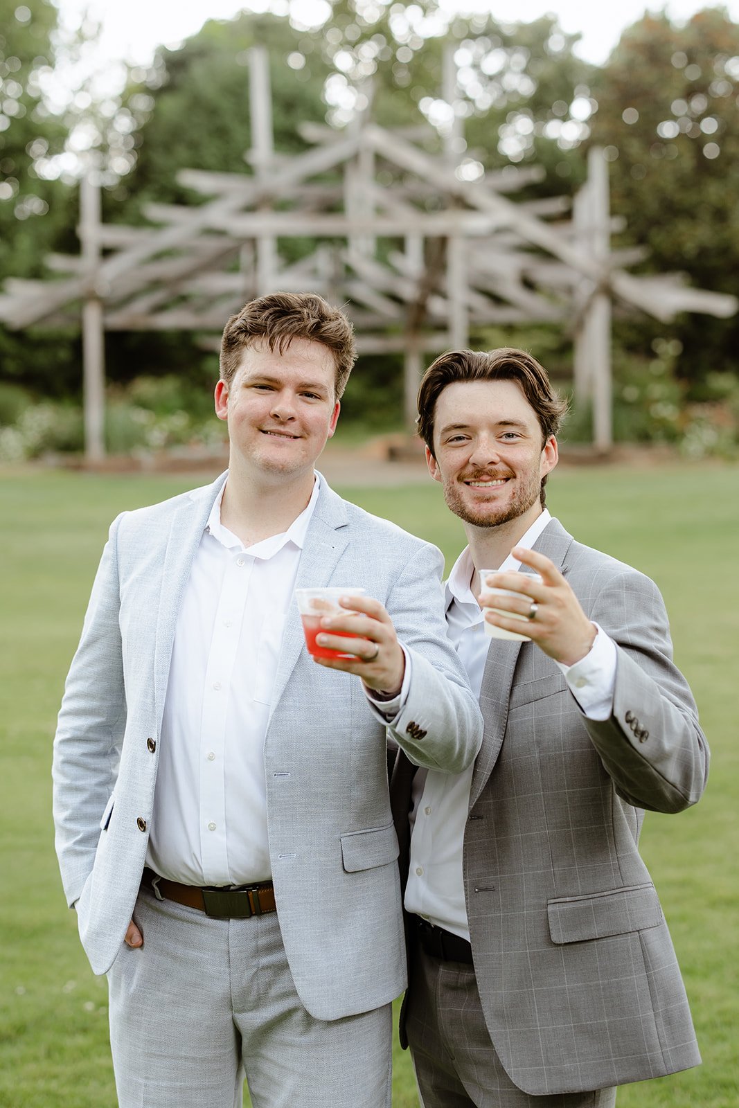 Two men in suits smiling and holding drinks at an outdoor event, with a wooden playground structure and trees in the background.