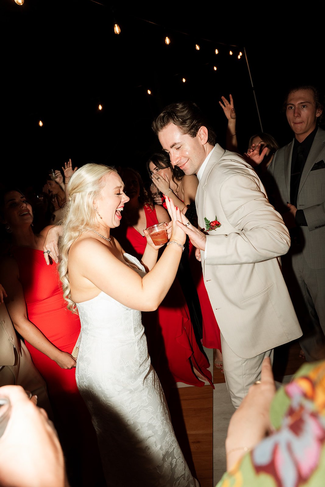A joyful bride and groom dancing and smiling at their wedding reception, surrounded by guests, under string lights at night.