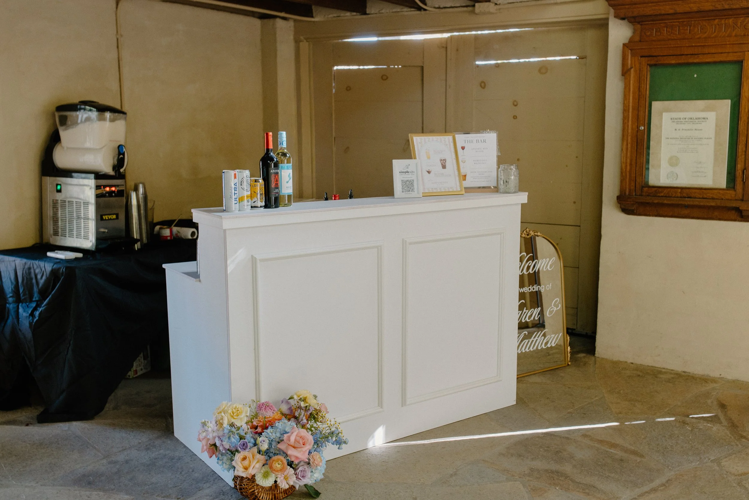 A white bar counter with bottles of wine, a napkin holder, and a promotional sign, next to a flower arrangement on the floor, in a rustic indoor setting with beige walls and a framed document on the wall.