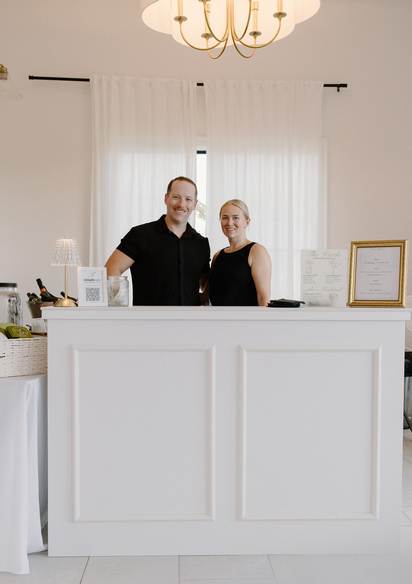A smiling man and woman standing behind a white reception desk in a well-lit room with white curtains and a chandelier overhead.
