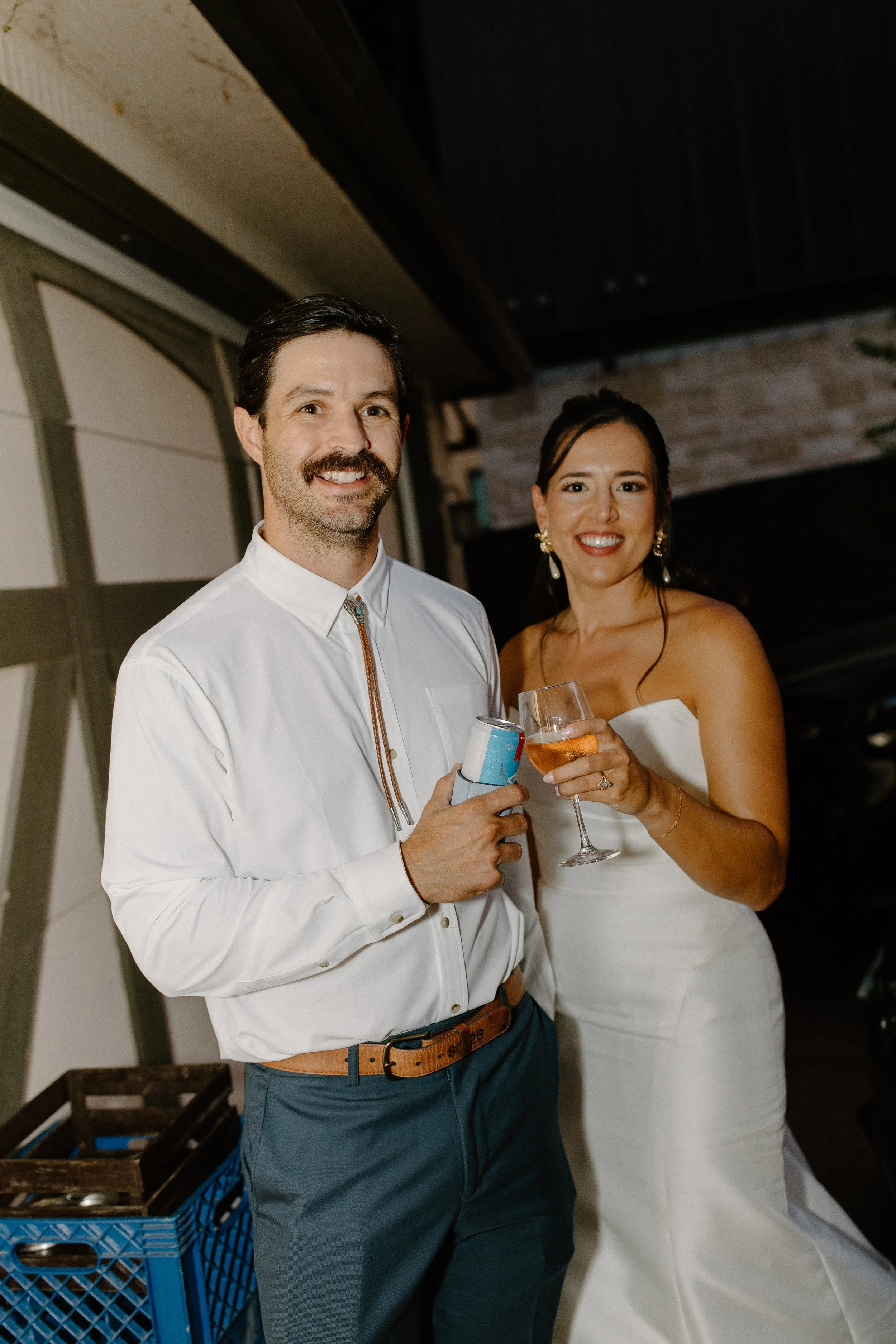 A man and a woman standing together at a social event, both holding drinks and smiling at the camera. The man is wearing a white shirt and dark pants, while the woman is in a strapless white dress with earrings. They are inside a warmly lit room with brick and wood decor.