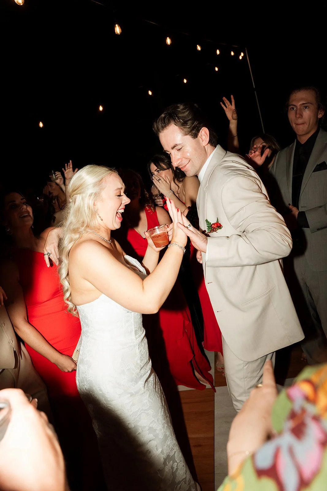 A bride and groom dancing and smiling at their wedding reception, surrounded by guests, with string lights overhead.