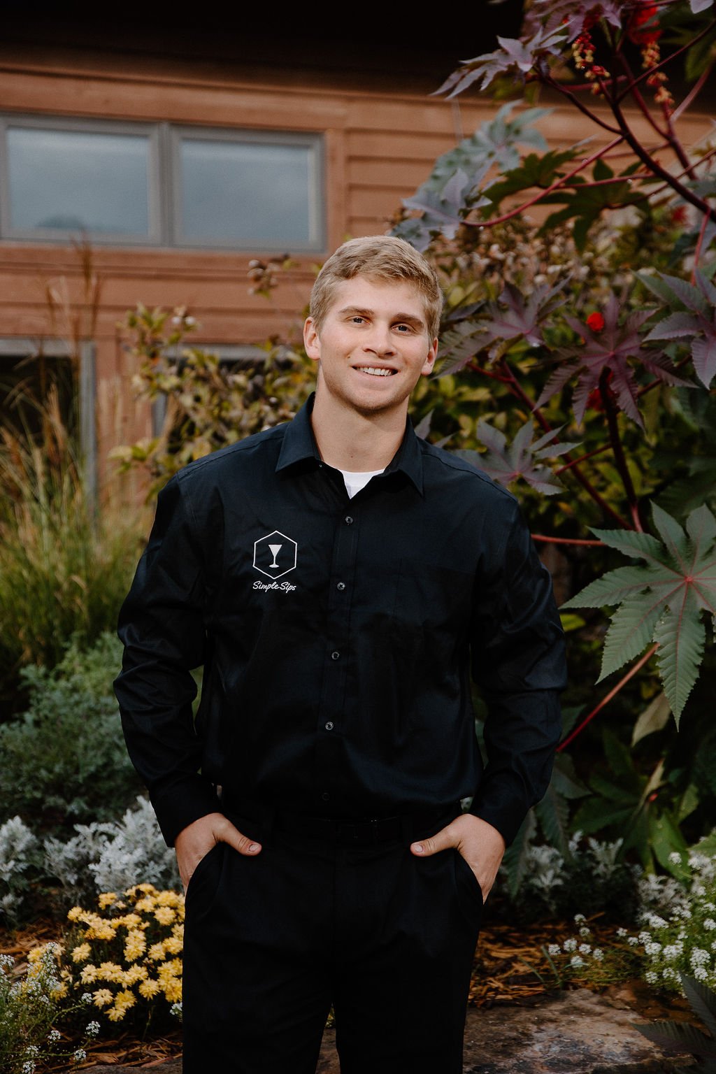 Young man in black dress shirt with logo, standing outdoors in front of a garden with colorful plants and a wooden house.