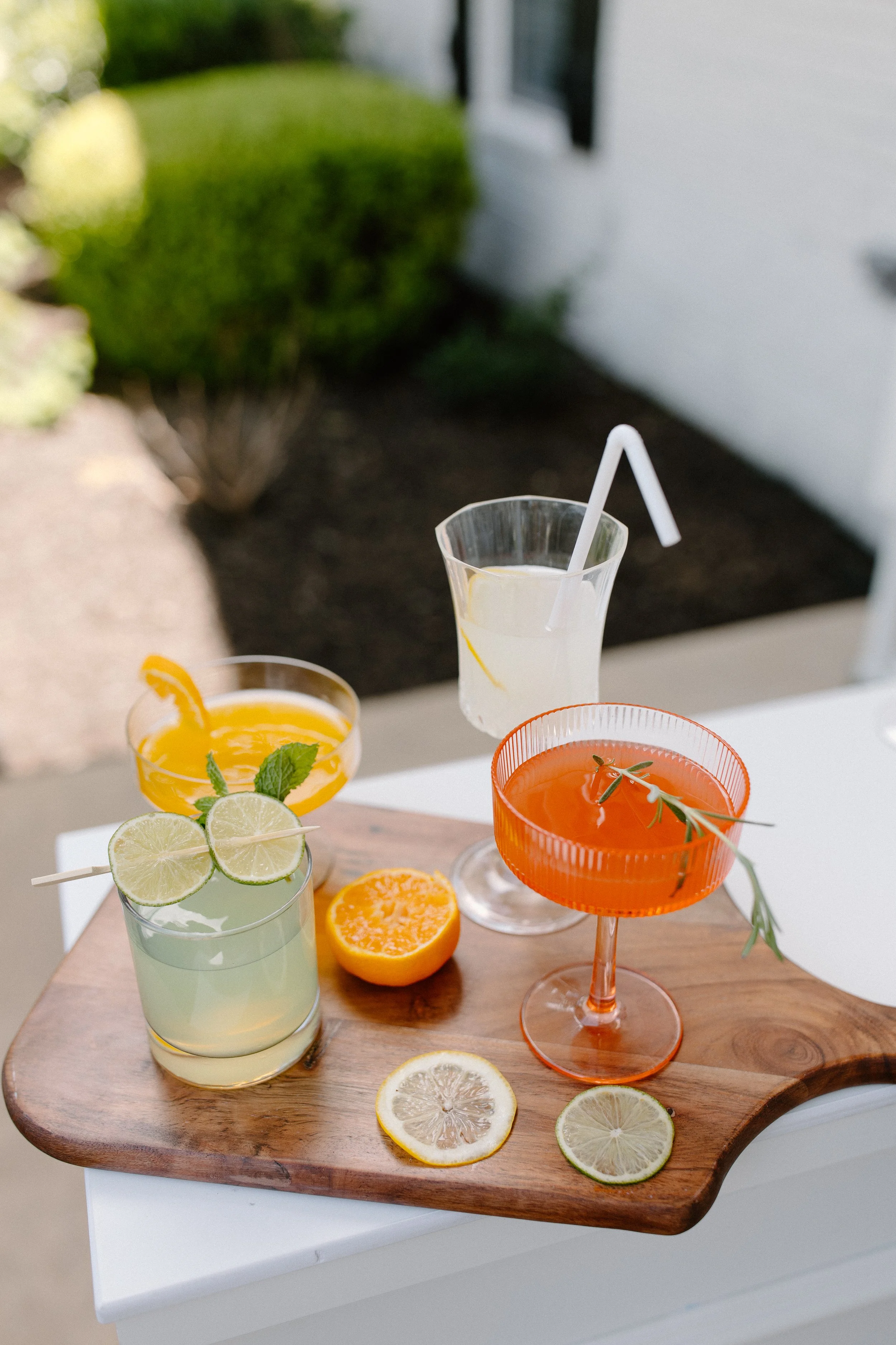 Colorful drinks with slices of citrus fruits and mint leaves on a wooden serving board, outdoors with blurred greenery and house in the background.