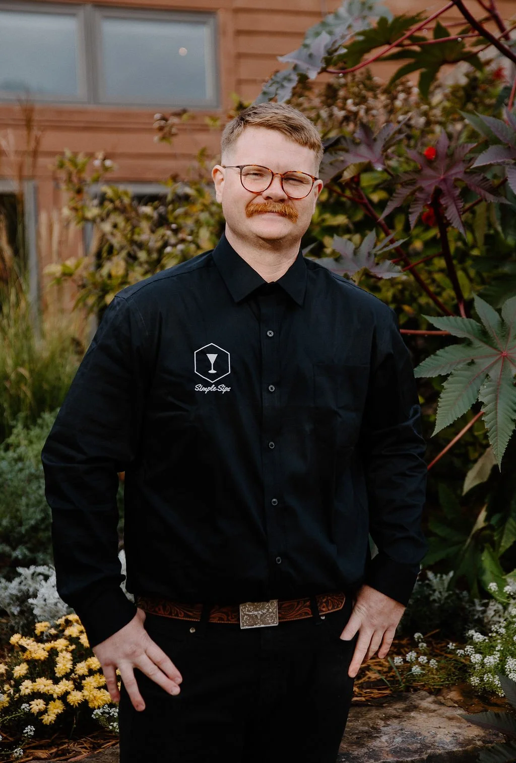 A man with glasses and a mustache wearing a black shirt with a logo on it, standing outdoors in front of plants and a wooden house.