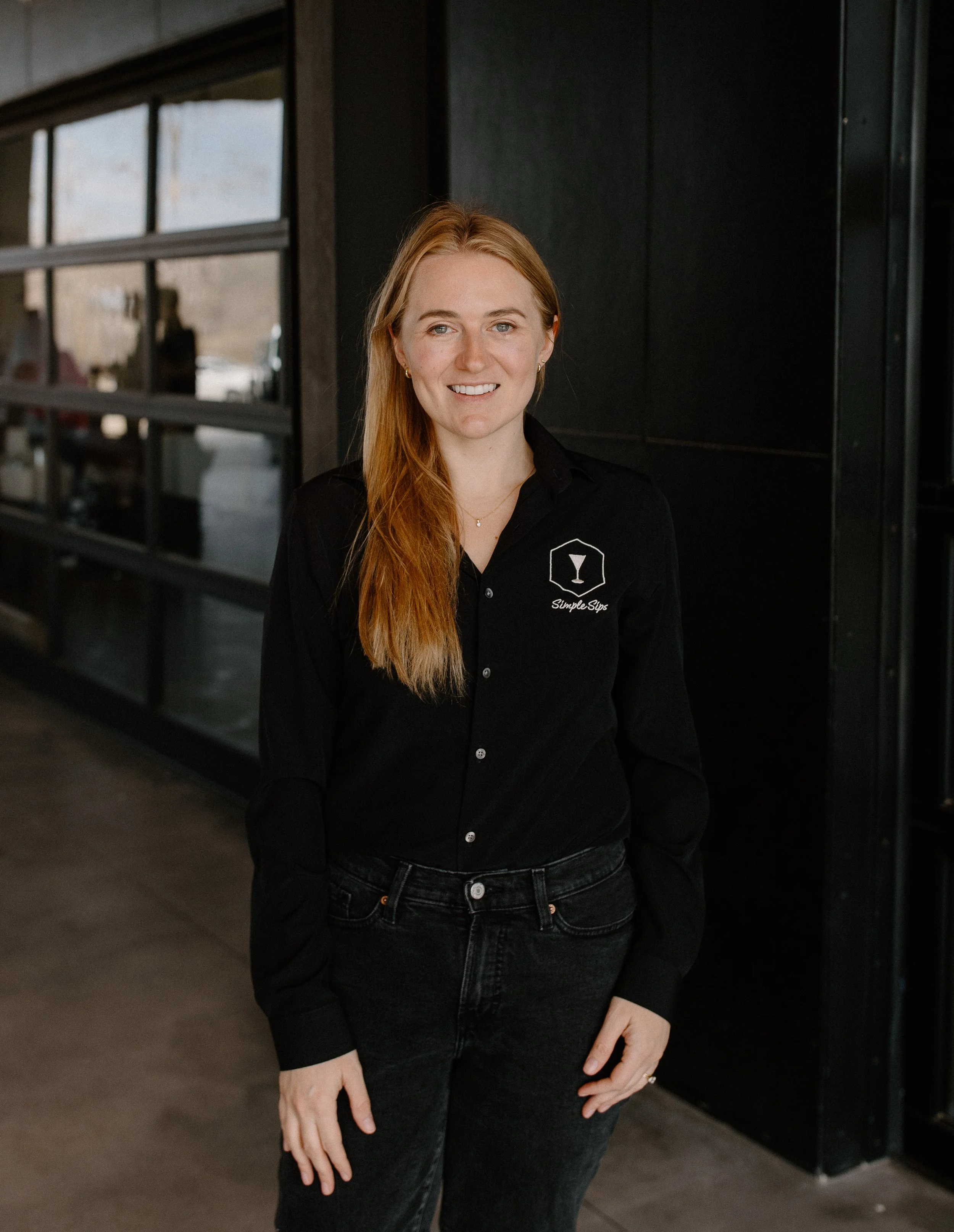 A woman standing indoors near a black wall, wearing a black button-up shirt with a logo of a cocktail glass and the text "SimpleSip" on it.