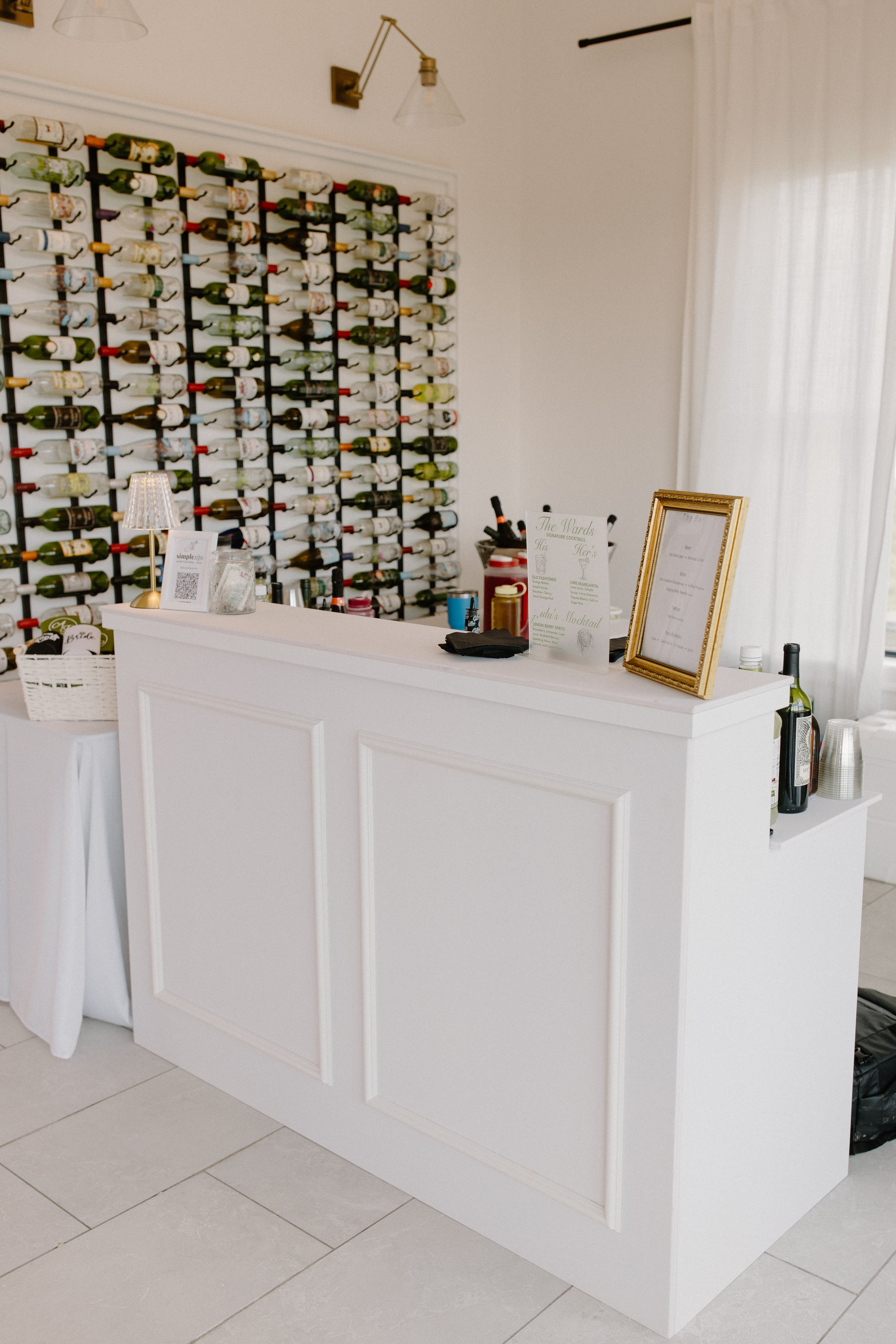 A white bar counter with a framed menu, some liquor bottles, and a small lamp, in front of a wall with a large wine bottle rack and white curtains allowing natural light to enter.