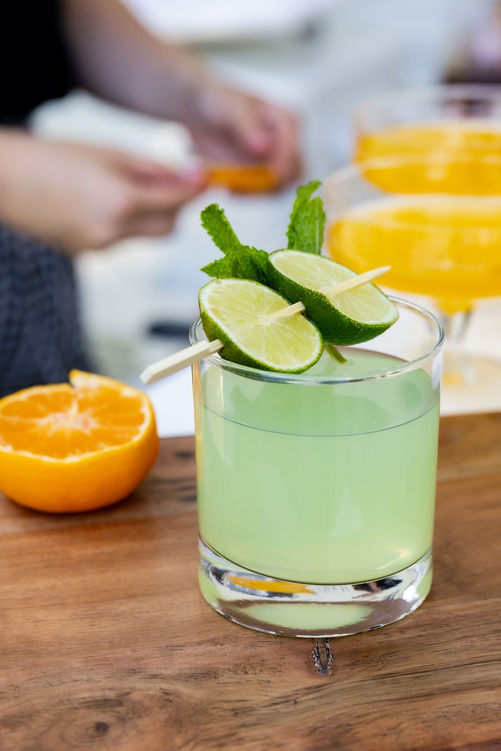 A green cocktail garnished with lime slices and mint leaves on a wooden surface, with an orange half and yellow drinks in the background.