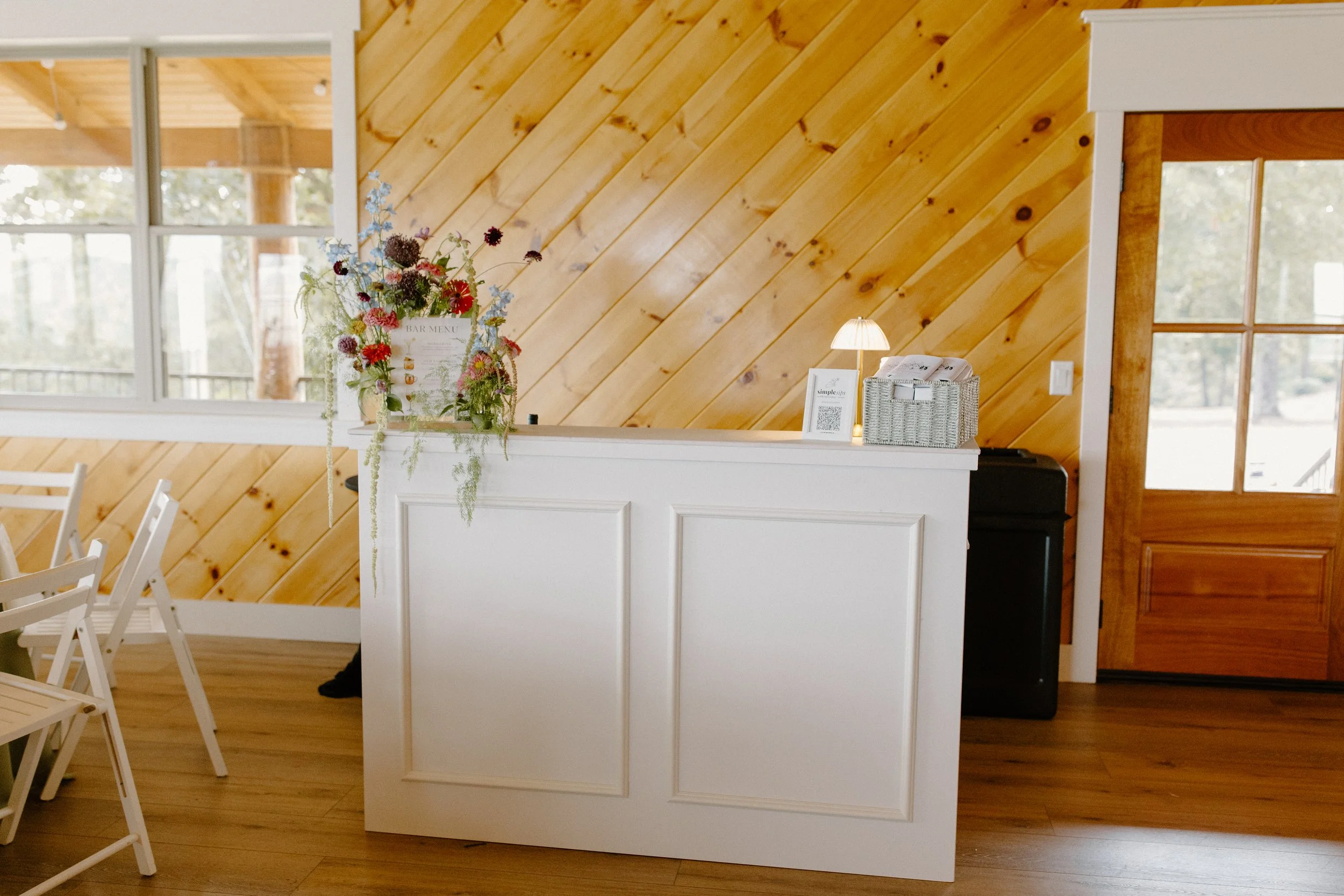 Interior of a room with wooden walls, white chairs, and a white counter with a floral arrangement and a lamp.