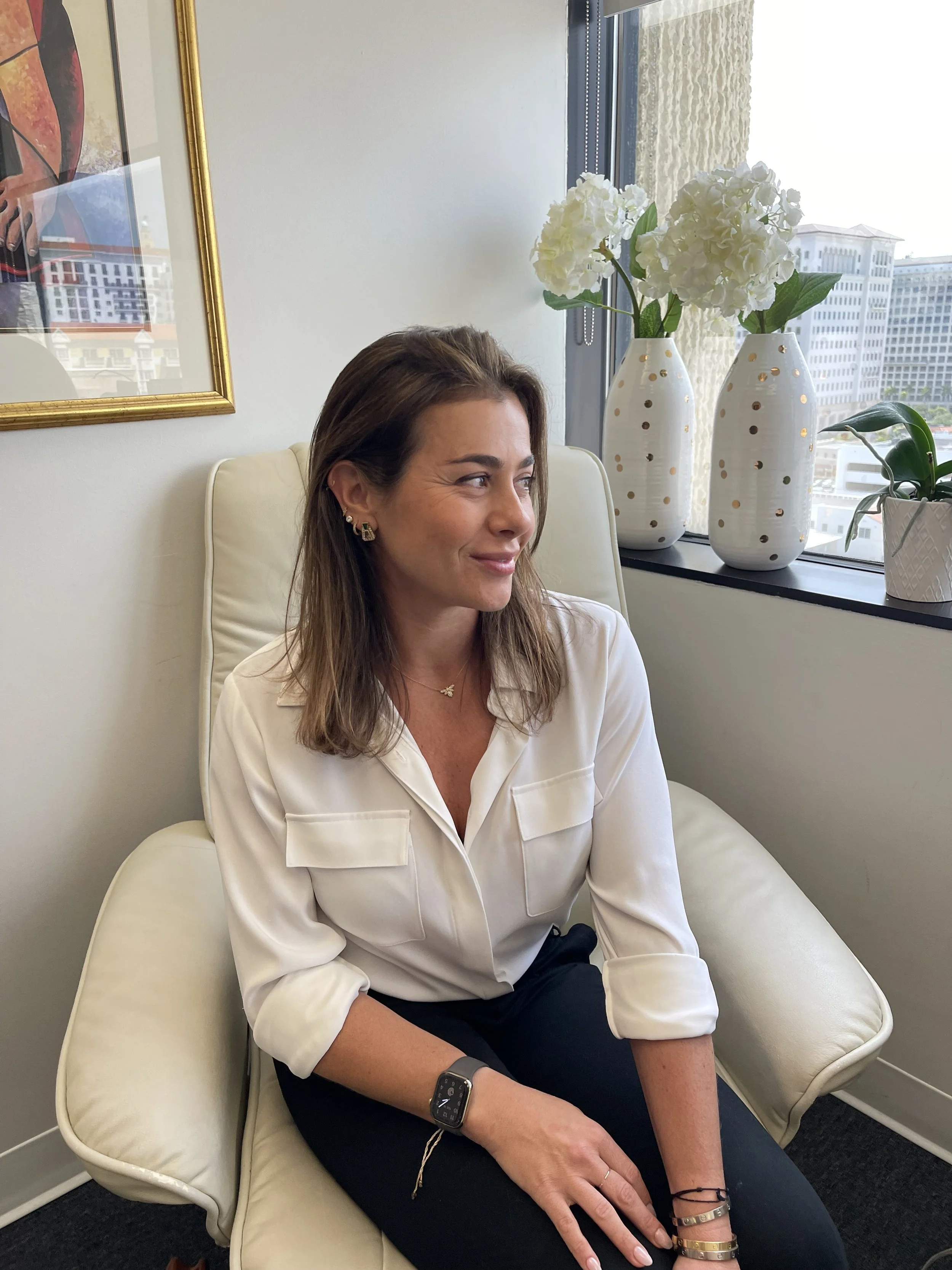 A woman with brown hair and earrings, wearing a white blouse, sitting on a cream-colored chair near a window. There are two white vases with white flowers and a smaller potted plant on the windowsill, with a cityscape visible outside.
