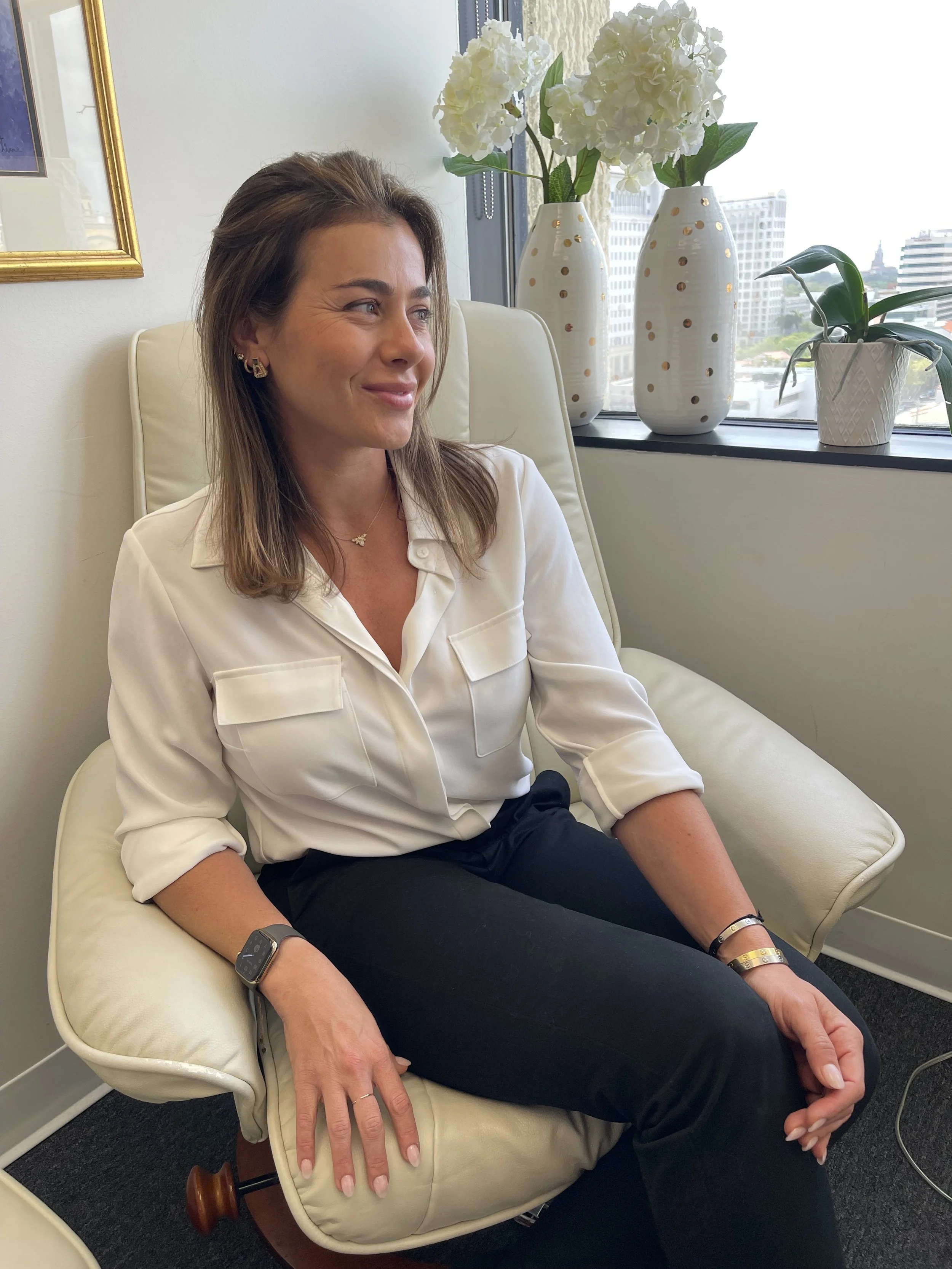 A cis-gendered, caucasian woman with shoulder-length brown hair, wearing a white blouse and black pants, sitting on a cream-colored chair in an office setting with a window behind her.