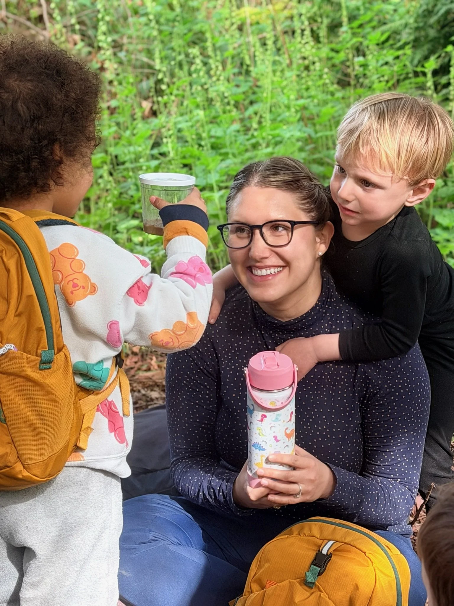 A woman sitting outdoors surrounded by children in a wooded area, holding a reusable water bottle with a pink lid, while one child offers her a cup of water from a container.