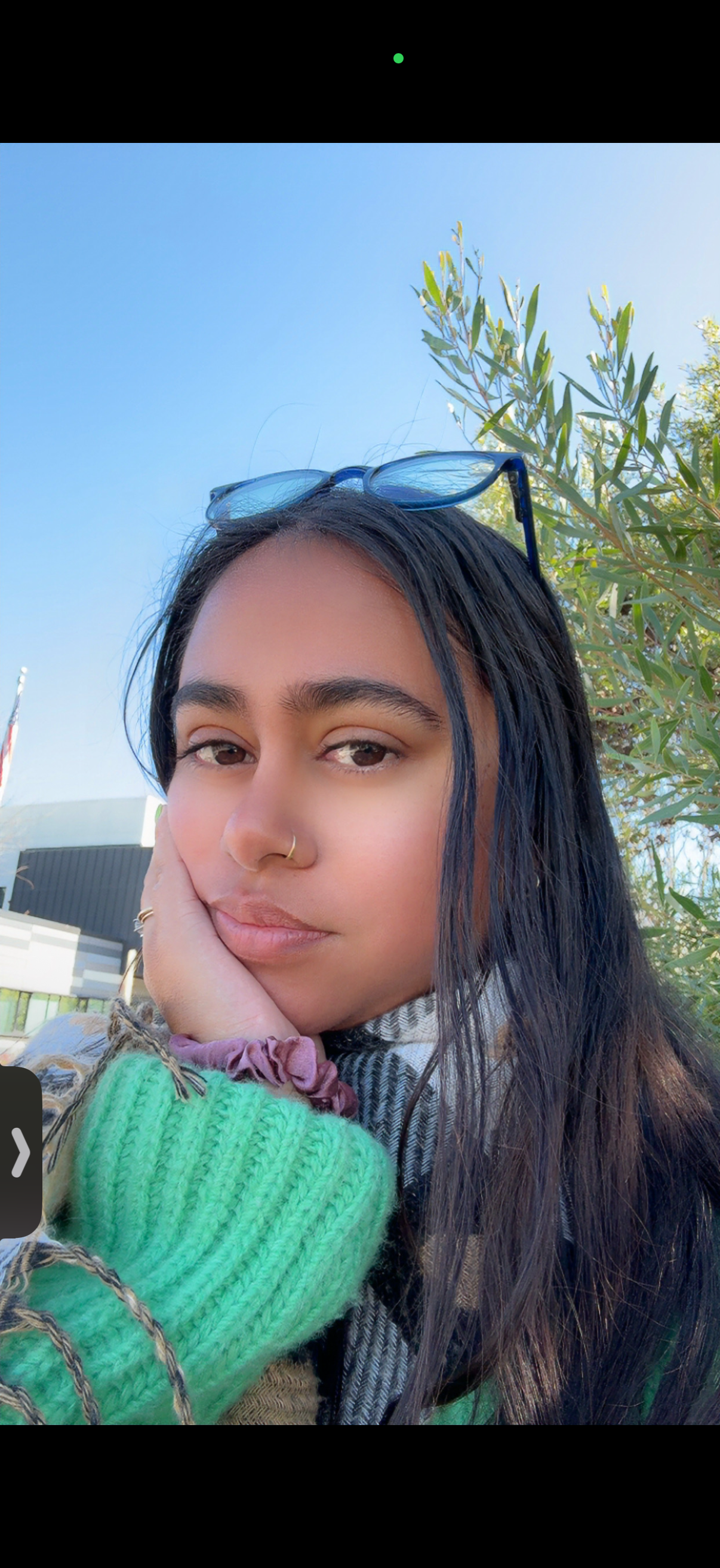 Close-up of a young woman with long dark hair, wearing sunglasses on her head, a nose ring, a green knitted sleeve, and a colorful outfit, resting her face on her hand outdoors with a blue sky, green leaves, and a building in the background.