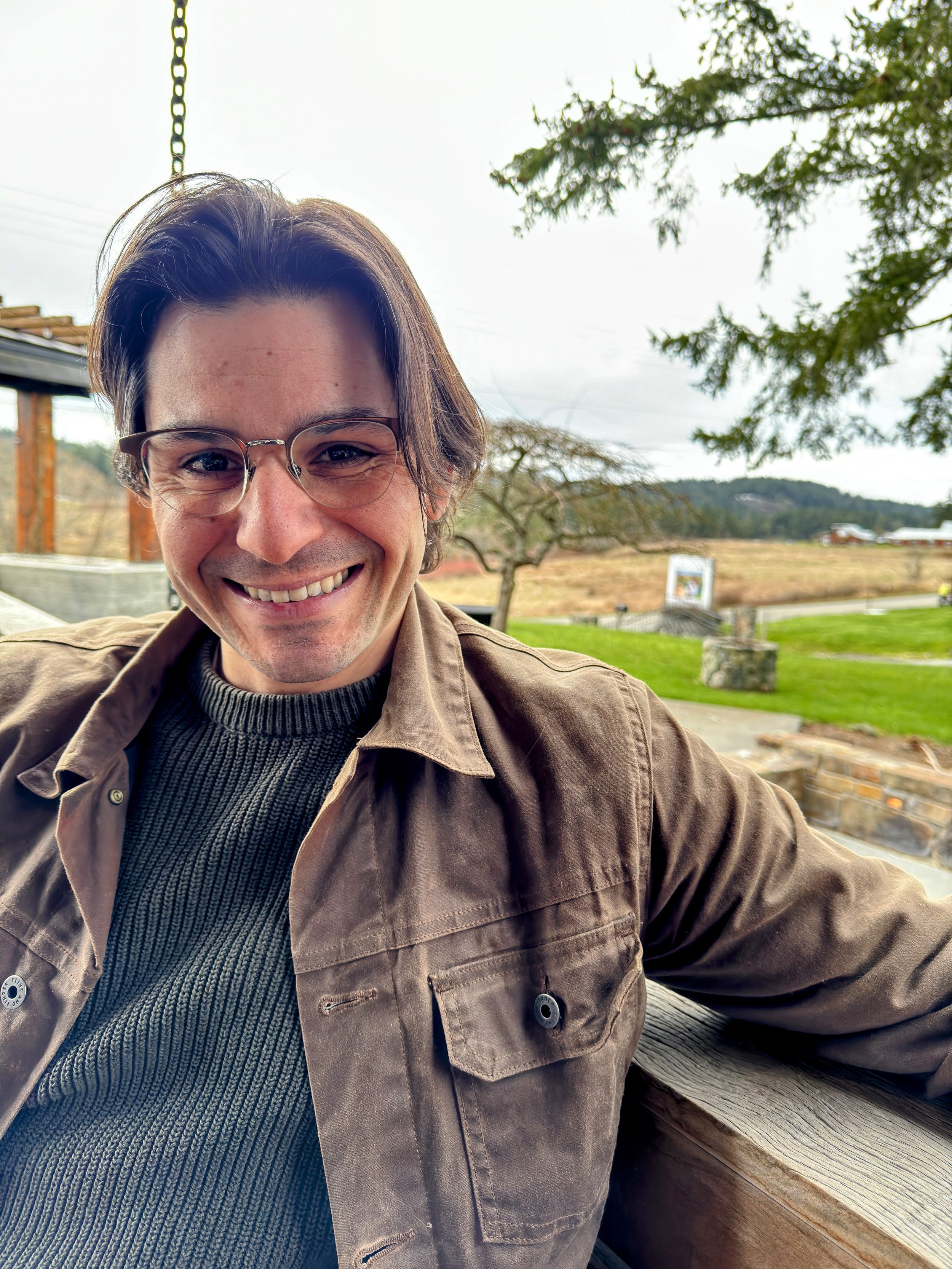 A smiling man with glasses wearing a tan sweatshirt with an REI logo, leaning on a railing in an indoor setting.