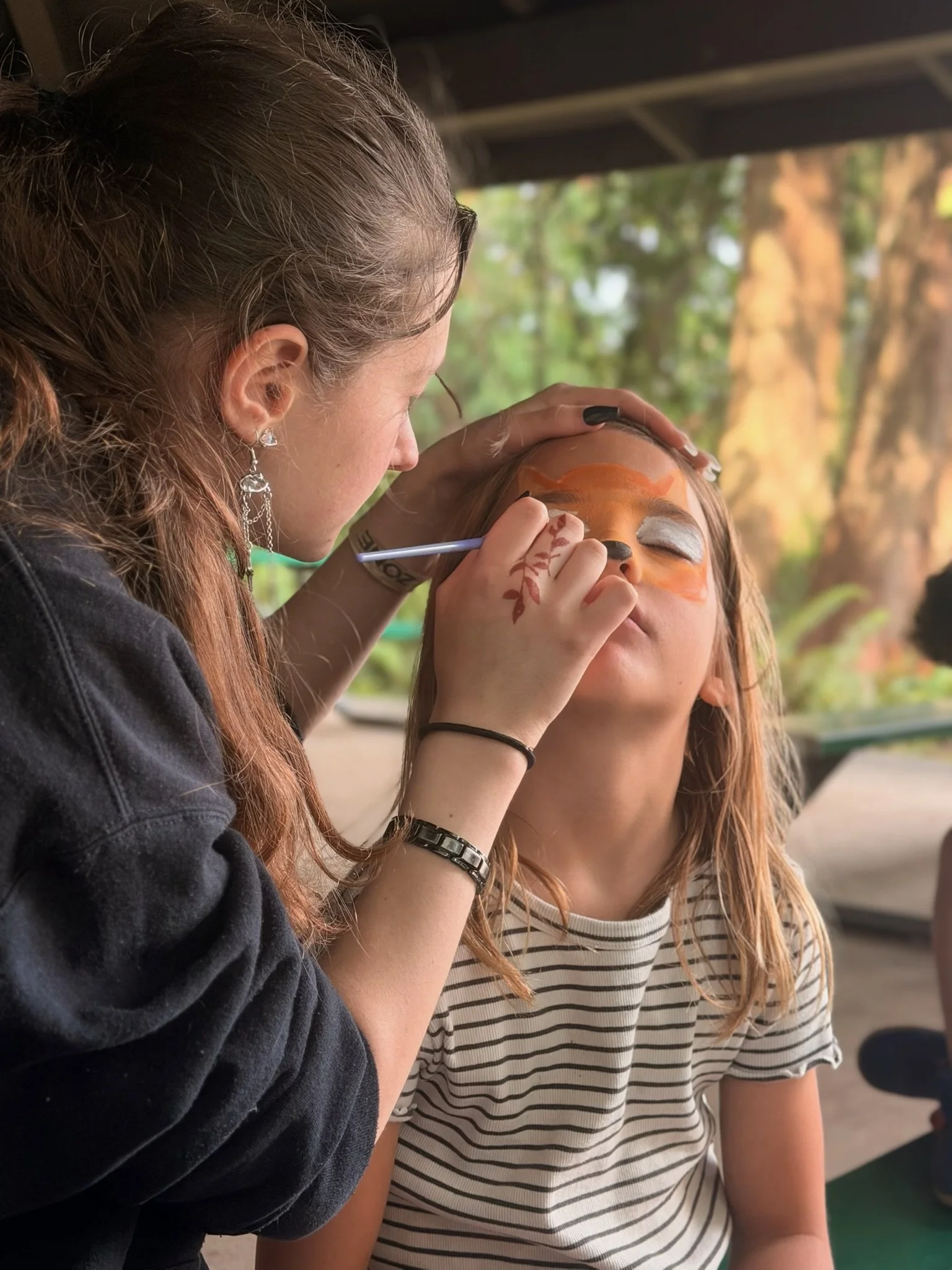 A woman with red hair, wearing a striped shirt, is having her face painted by another woman with earrings and a black jacket.