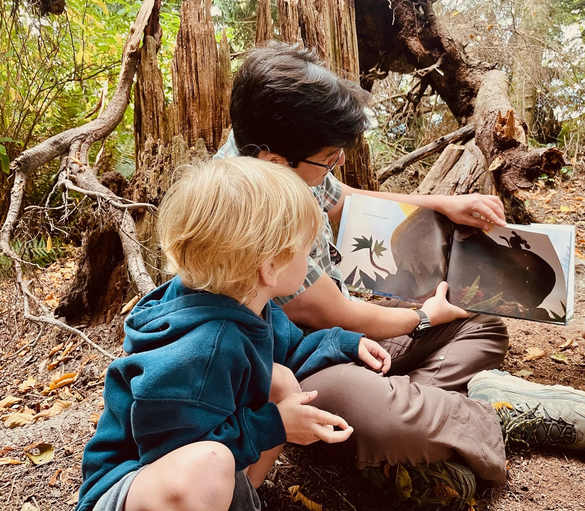 People sitting on the ground in a forest, reading a book about dinosaurs.