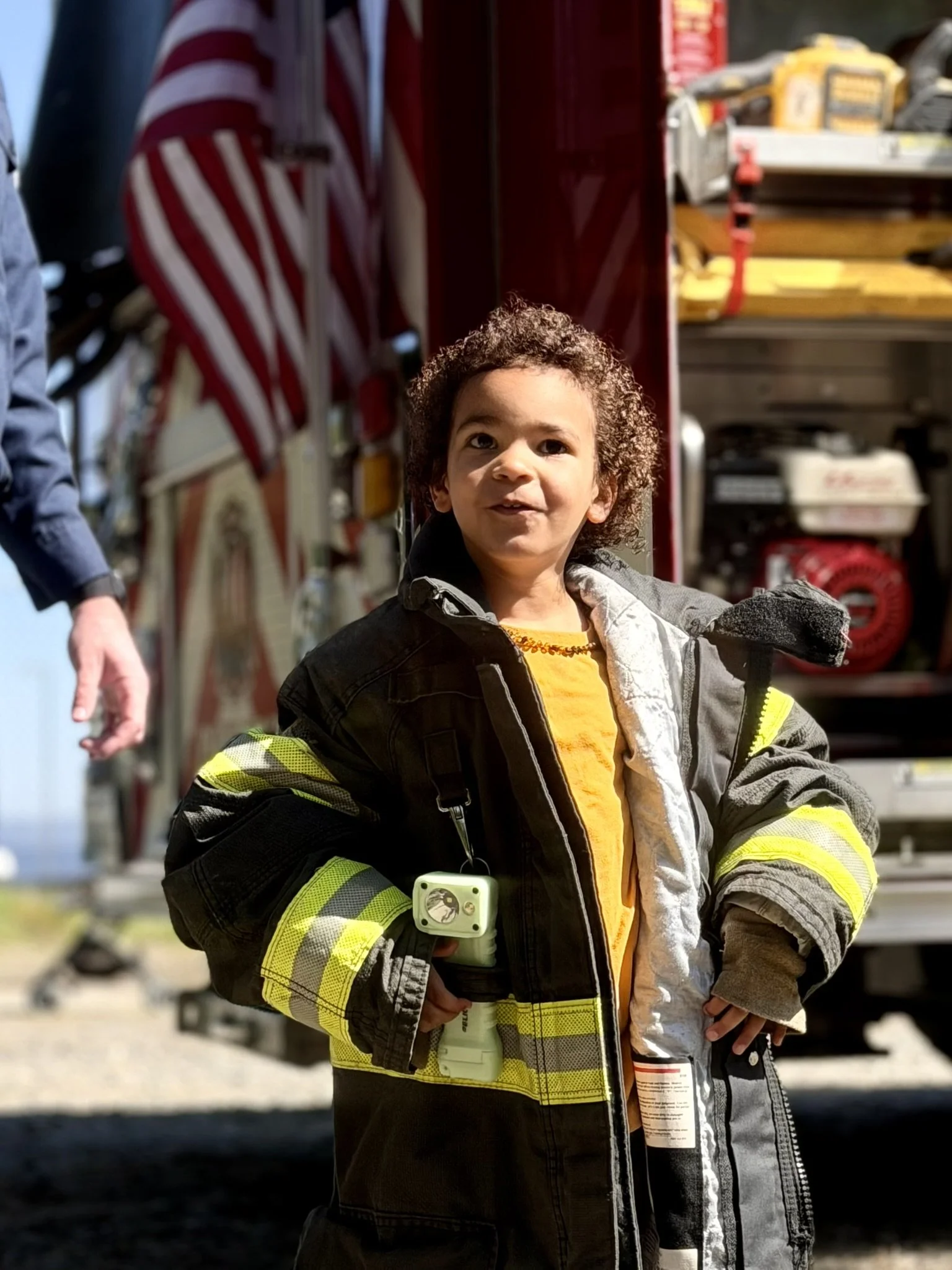 A young boy with curly hair wearing a firefighter uniform with reflective stripes, standing in front of a fire truck with firefighting equipment on a sunny day.