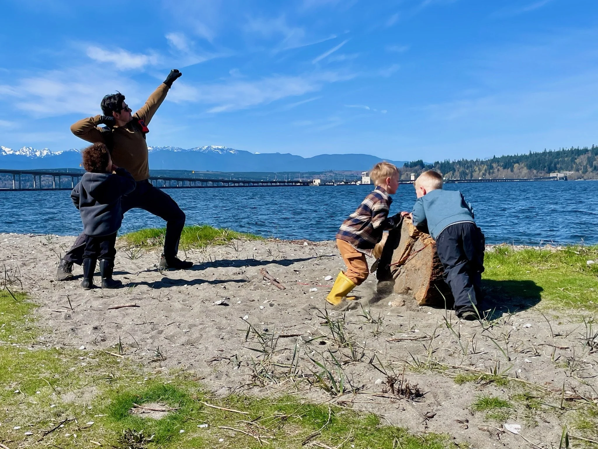 A woman and three children outdoors near a body of water with a bridge and snow-capped mountains in the background. The children are crouching and examining something in the sand, while the woman stands nearby, looking up and pointing.