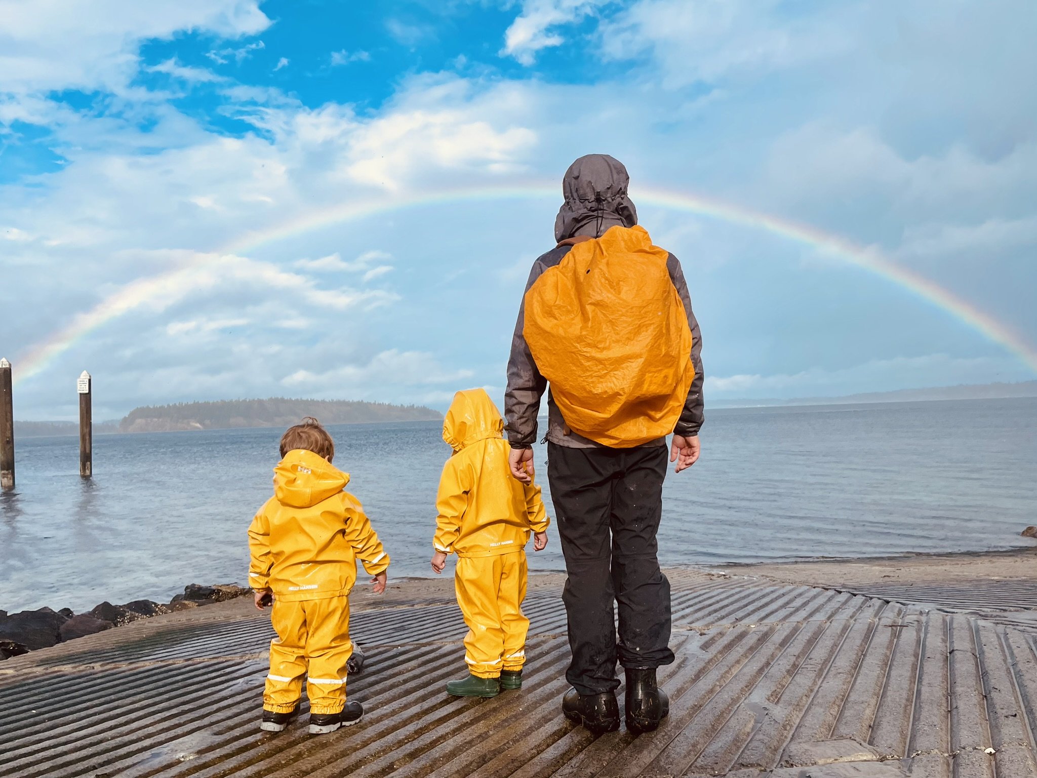 A person and two children in yellow raincoats and boots standing on a dock and looking at a rainbow over the water and distant land.