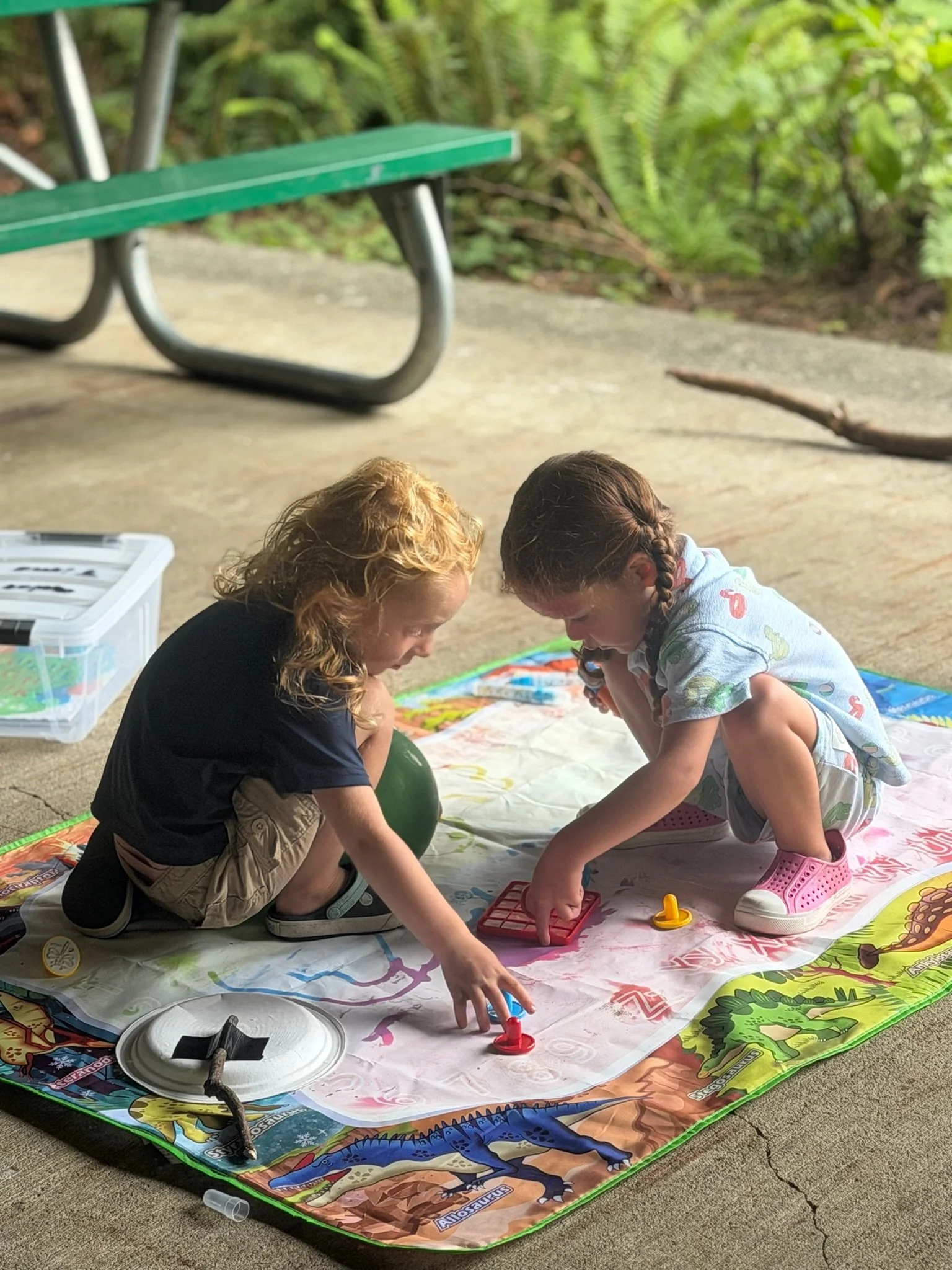 Two children playing a board game on a colorful mat outdoors, with a green park bench in the background and natural greenery surrounding them.