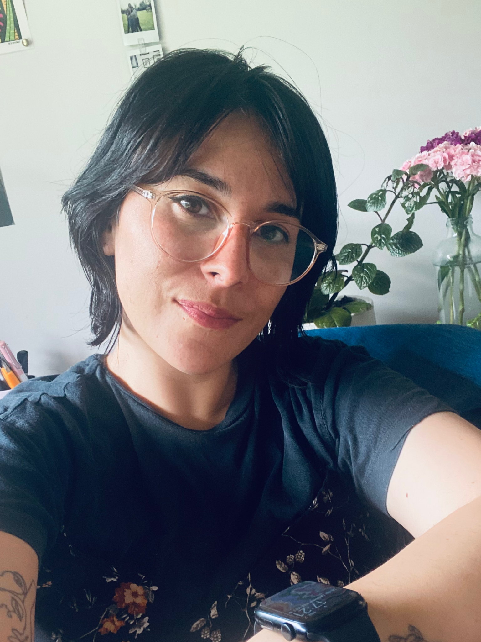 A woman with short black hair, wearing glasses, sitting at a table with a floral skirt, taking a selfie. In the background, there are pictures on the wall and a vase with pink and purple flowers.