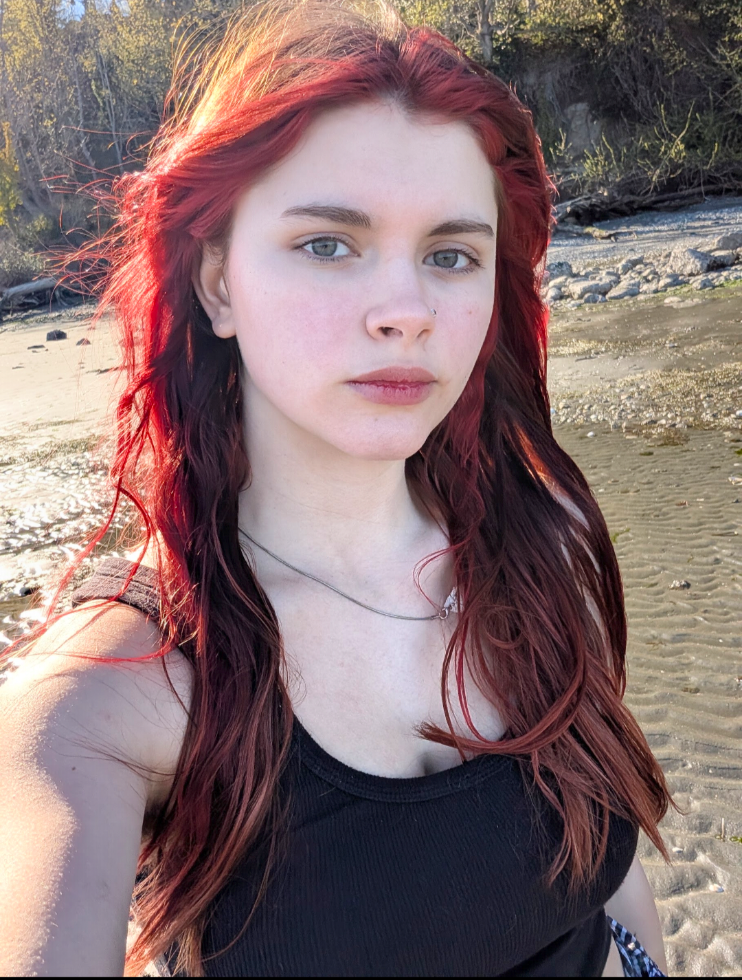 Young woman with long red hair and fair skin taking a selfie on a beach with sand and rocks, during sunset or late afternoon.