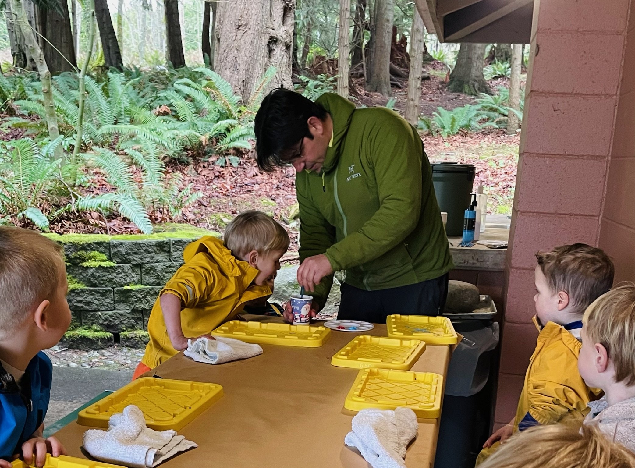 A group of children gathered around a table outdoors, engaged in a hands-on activity with an adult, with lush greenery and trees in the background.
