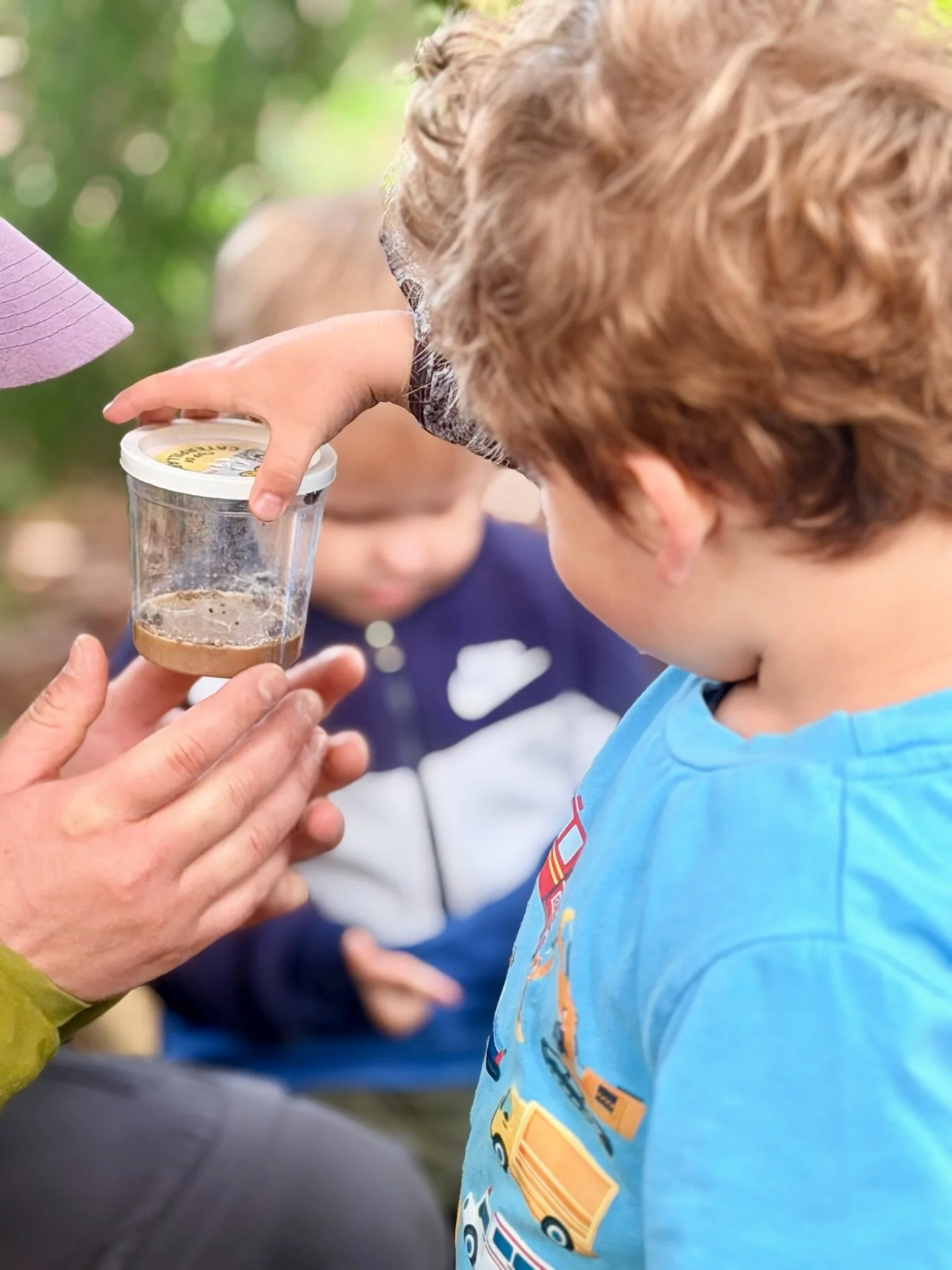 A young boy in a blue T-shirt with colorful cars on it receives a scoop of ice cream from an adult outdoors, with a blurred background of greenery.