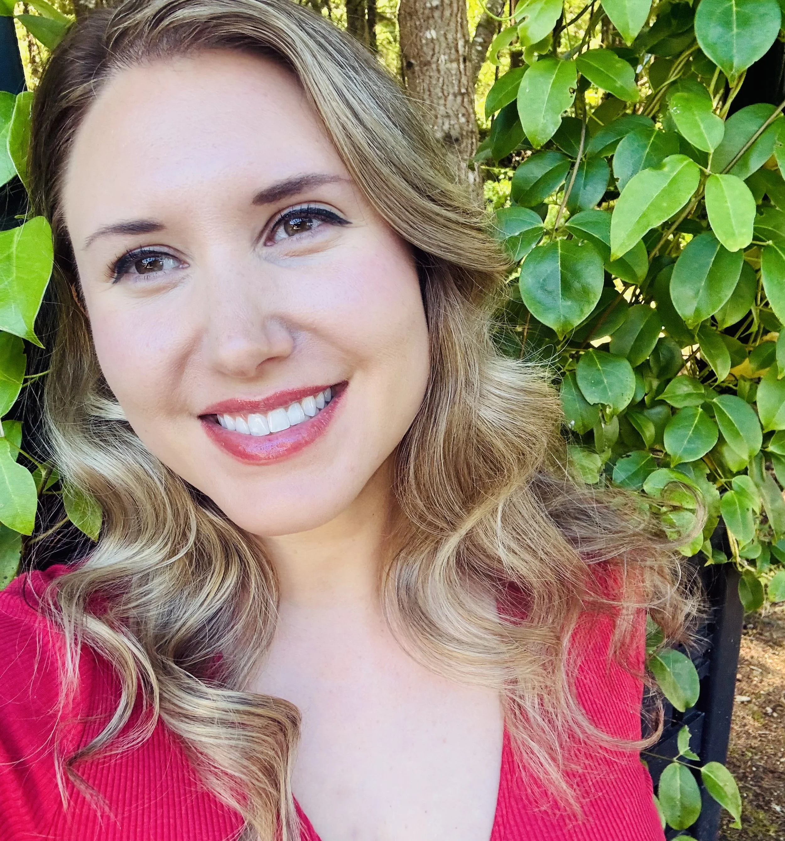 Close-up of a woman's face smiling outdoors, with green leaves and tree trunk in the background, wearing a red top and makeup.