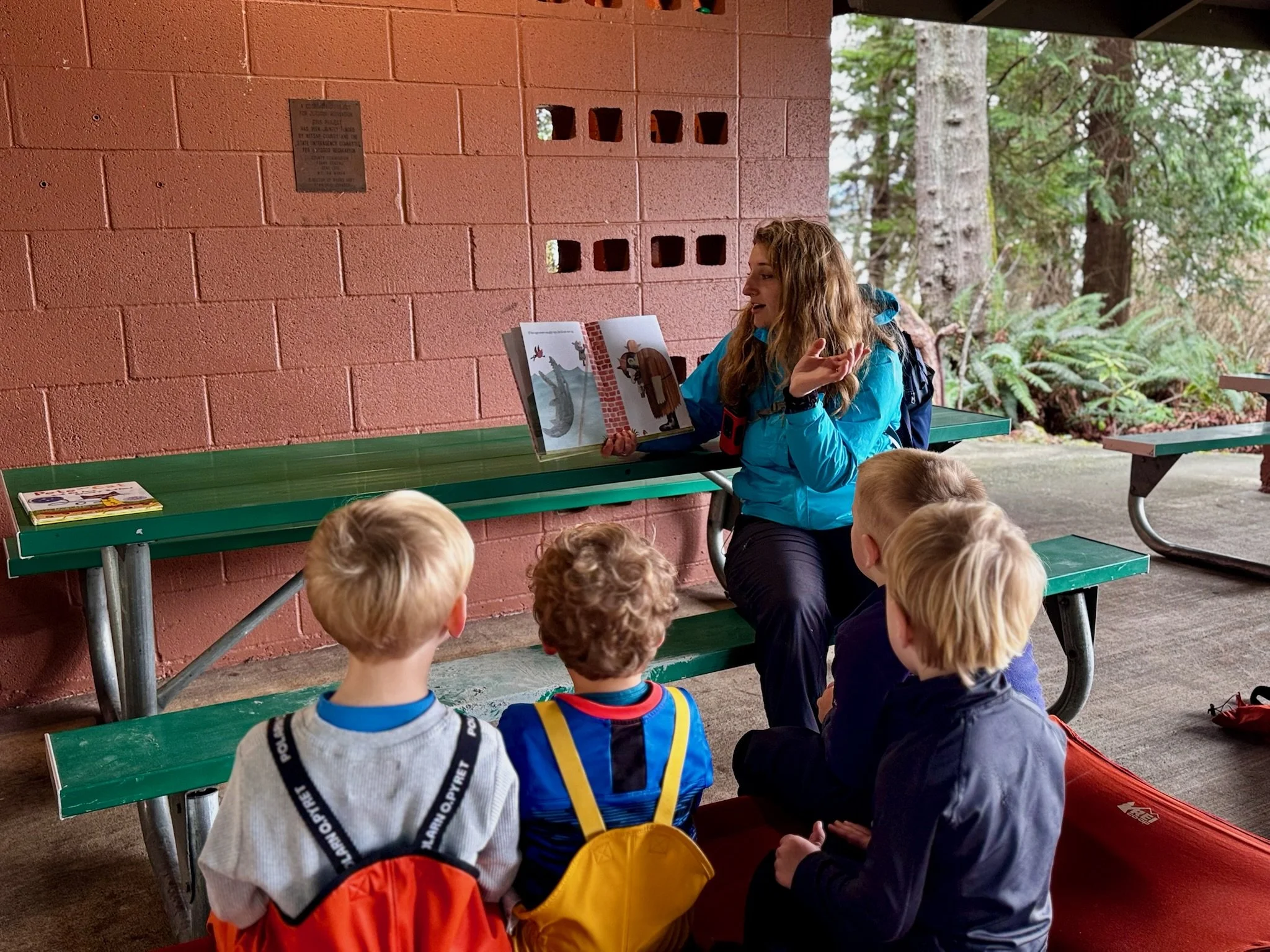 A woman reading a book to a group of children seated at a picnic table outdoors, surrounded by trees and greenery.