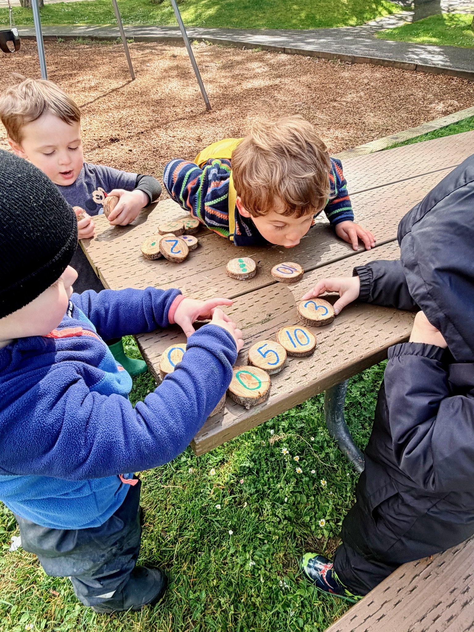 Children playing with wooden tokens labeled with numbers on a picnic table outdoors, with grass and a playground slide in the background.