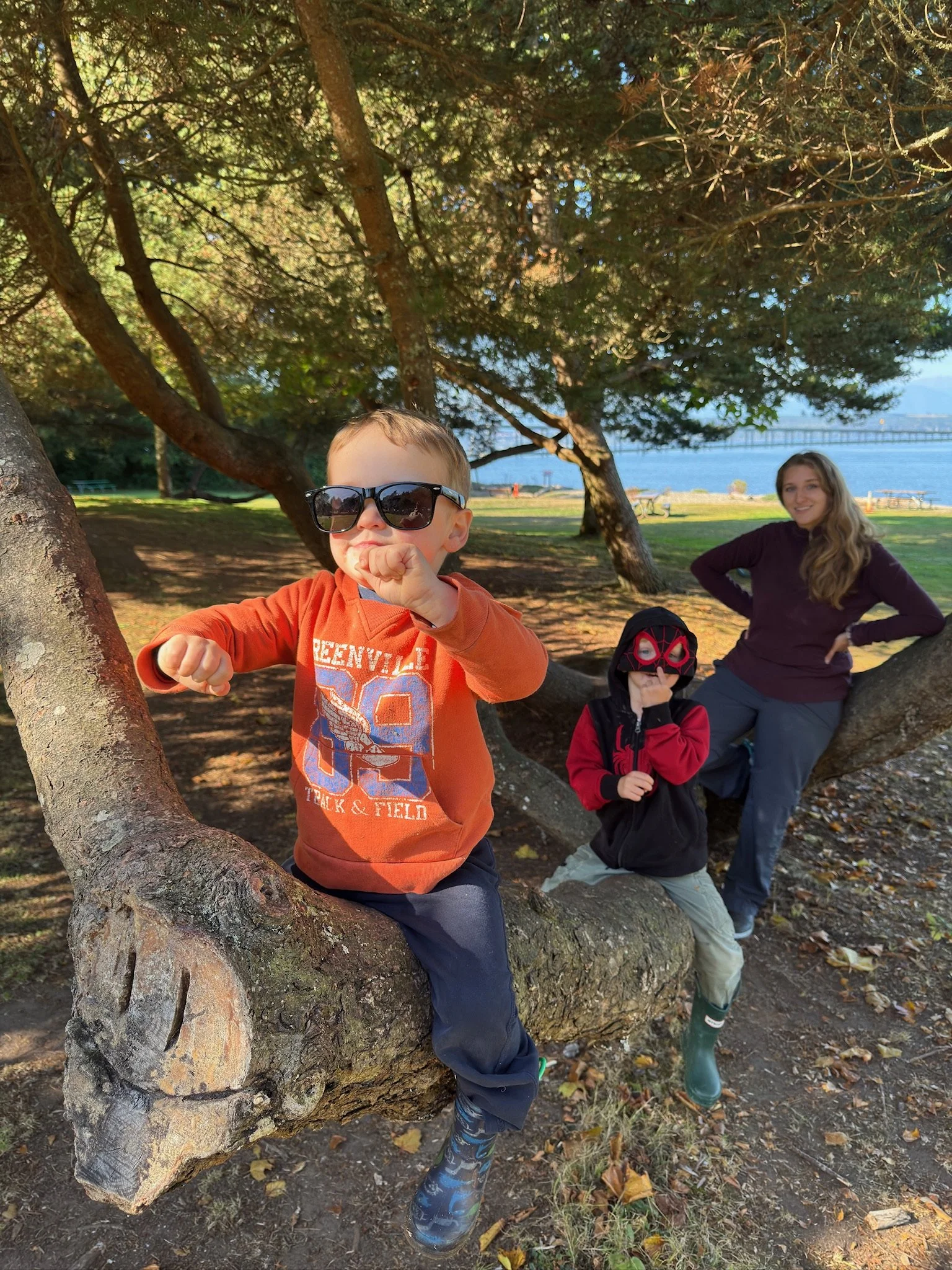 Four children are playing outdoors near a large tree with a view of a body of water and a bridge in the background. One child, wearing sunglasses and an orange sweatshirt, is sitting in the tree and flexing his arm. Another child, dressed in a red an