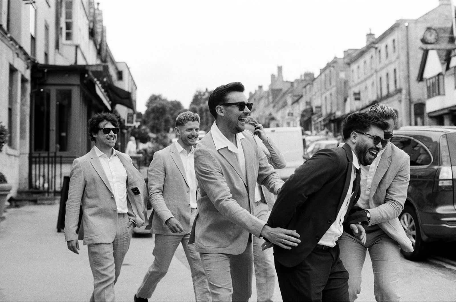 A groom and his groomsmen getting ready at The Bull in Burford for his wedding at Worton Kitchen Garden. All the guys are laughing and walking down the street outside the hotel.