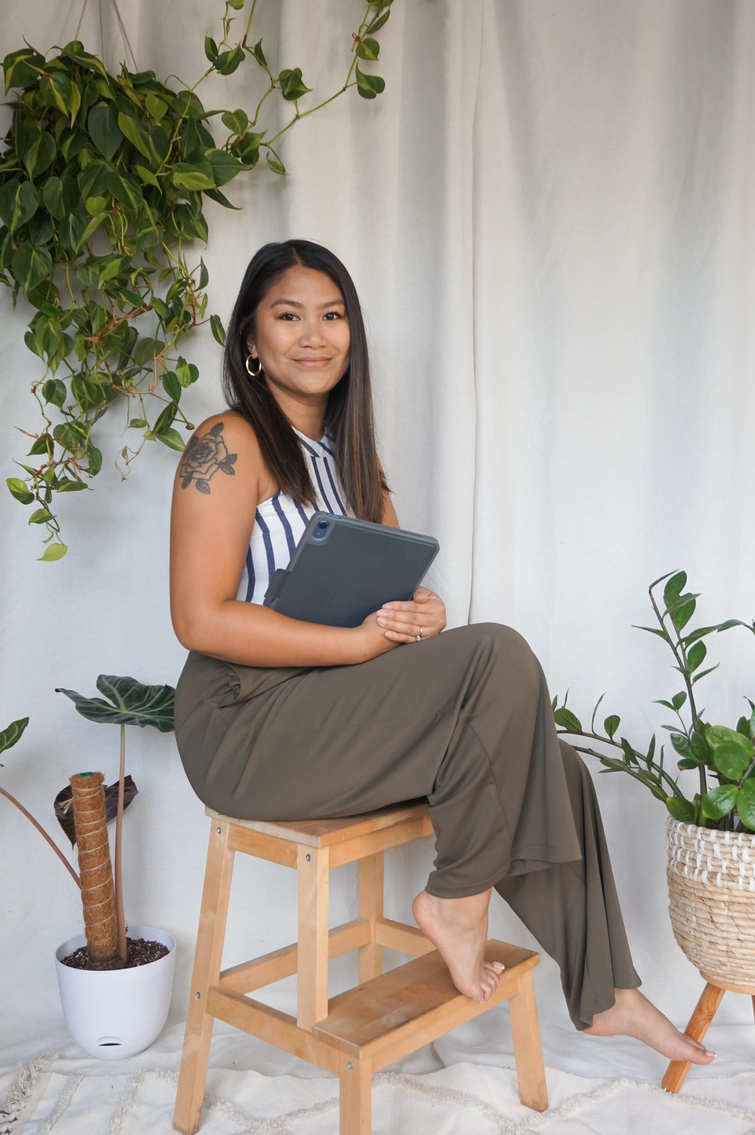 Christina Hellman, owner of Greenhaus Studio, sitting on a wooden stool, smiling at the camera while holding an ipad. The background is styled with plants and greenery.